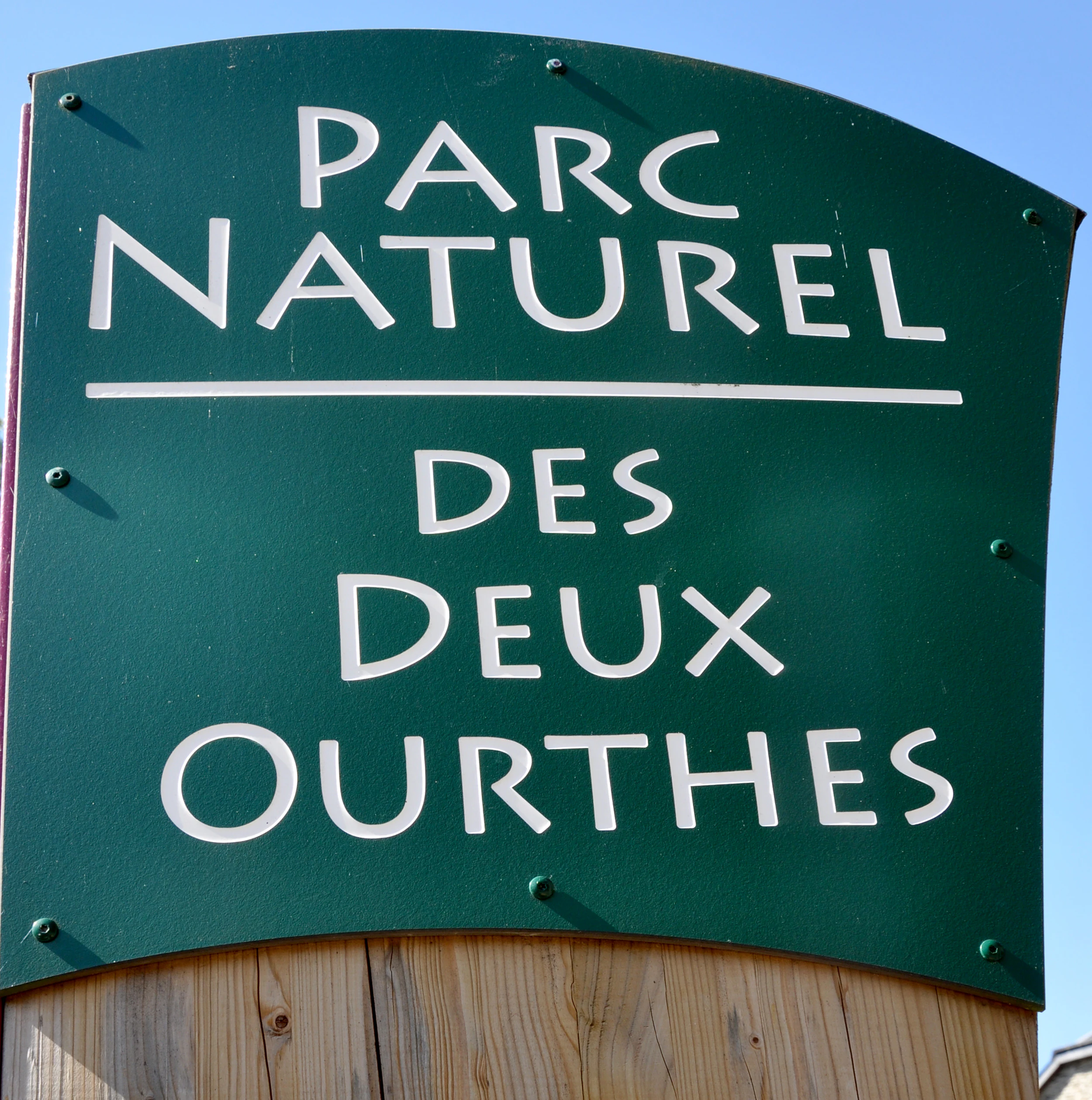A close-up shot of a dark green, arched park entrance sign with white text reading Parc Naturel des Deux Ourthes, mounted on a wooden post against a blue sky.