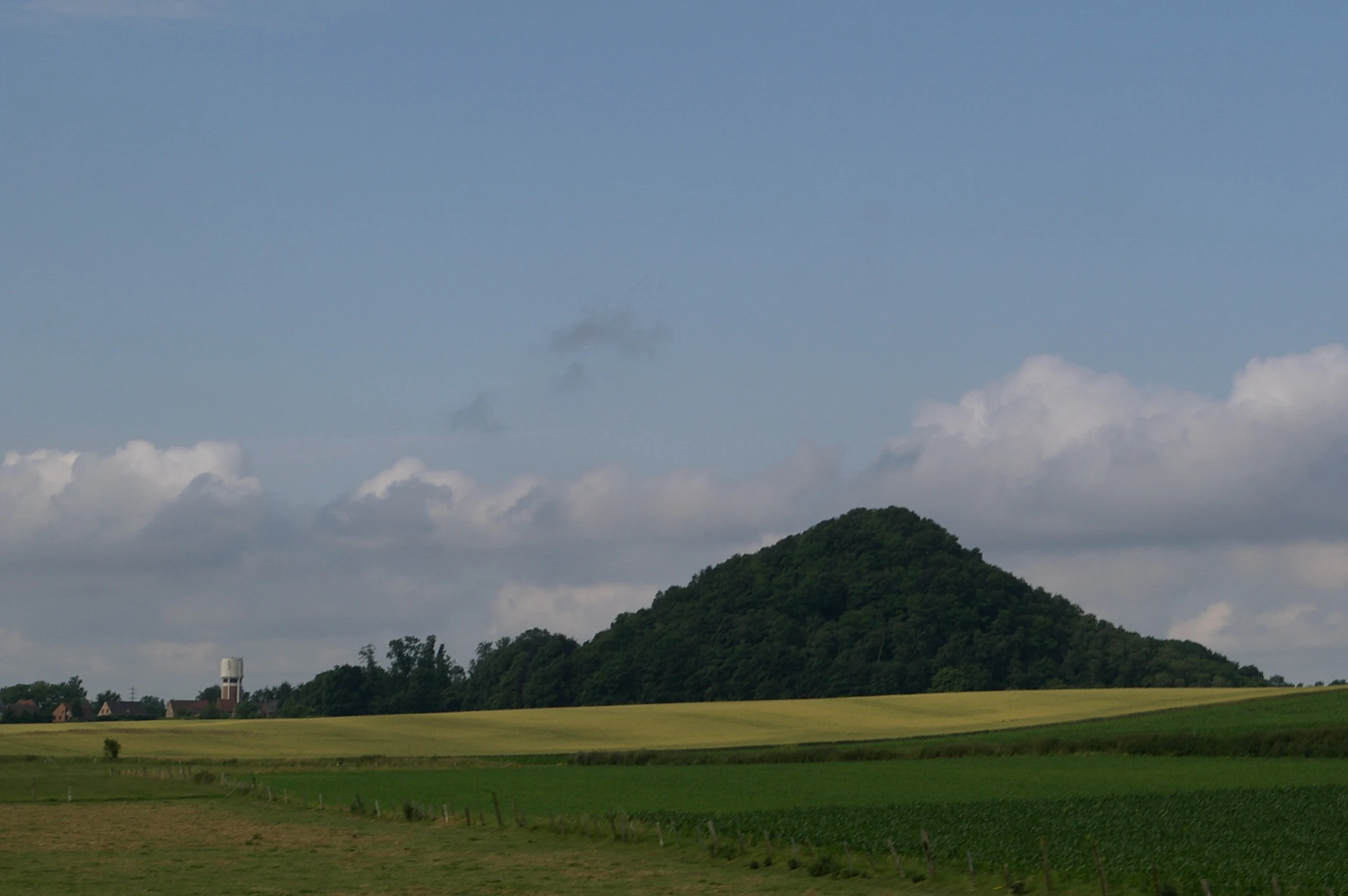 A conical, forest-covered spoil tip rising from green and yellow agricultural fields under a partly cloudy sky.
