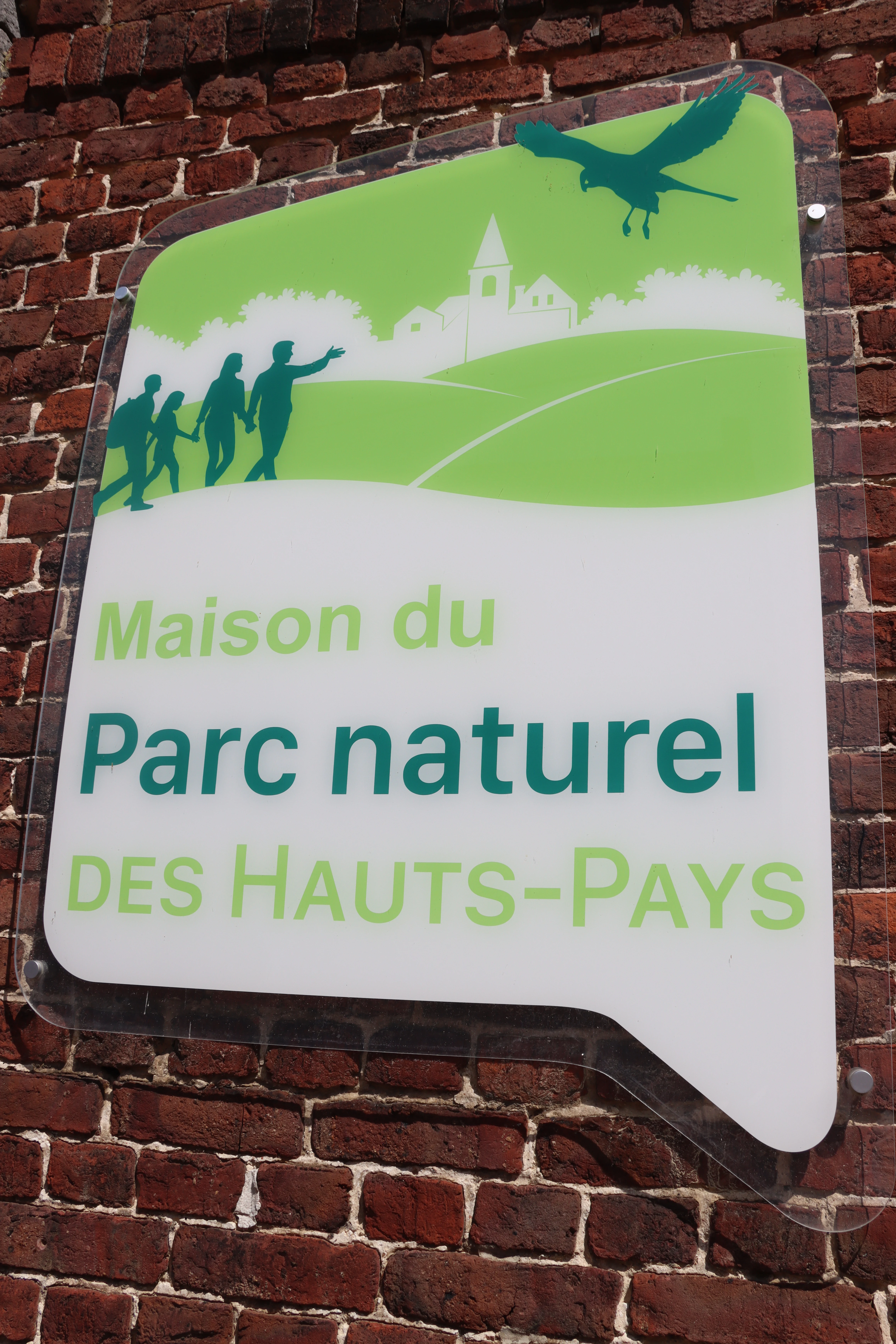 A sign for the Maison du Parc naturel des Hauts-Pays mounted on a red brick wall, featuring green graphics of people, a bird, and a church silhouette, with green and dark text.