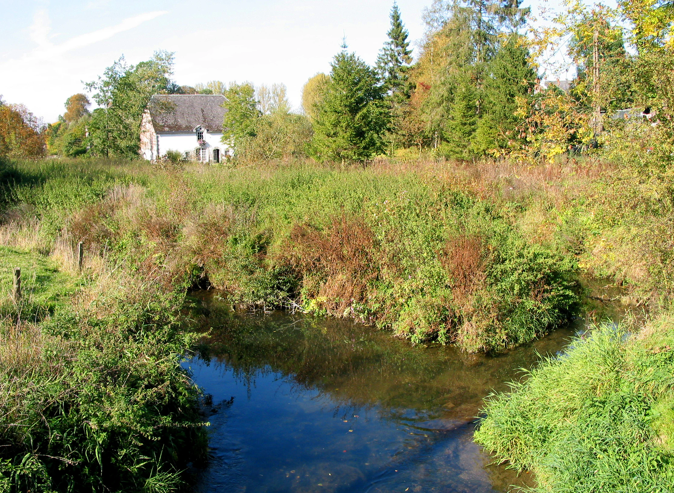 A peaceful stream winds through lush green vegetation and trees, with a stone house visible in the background.