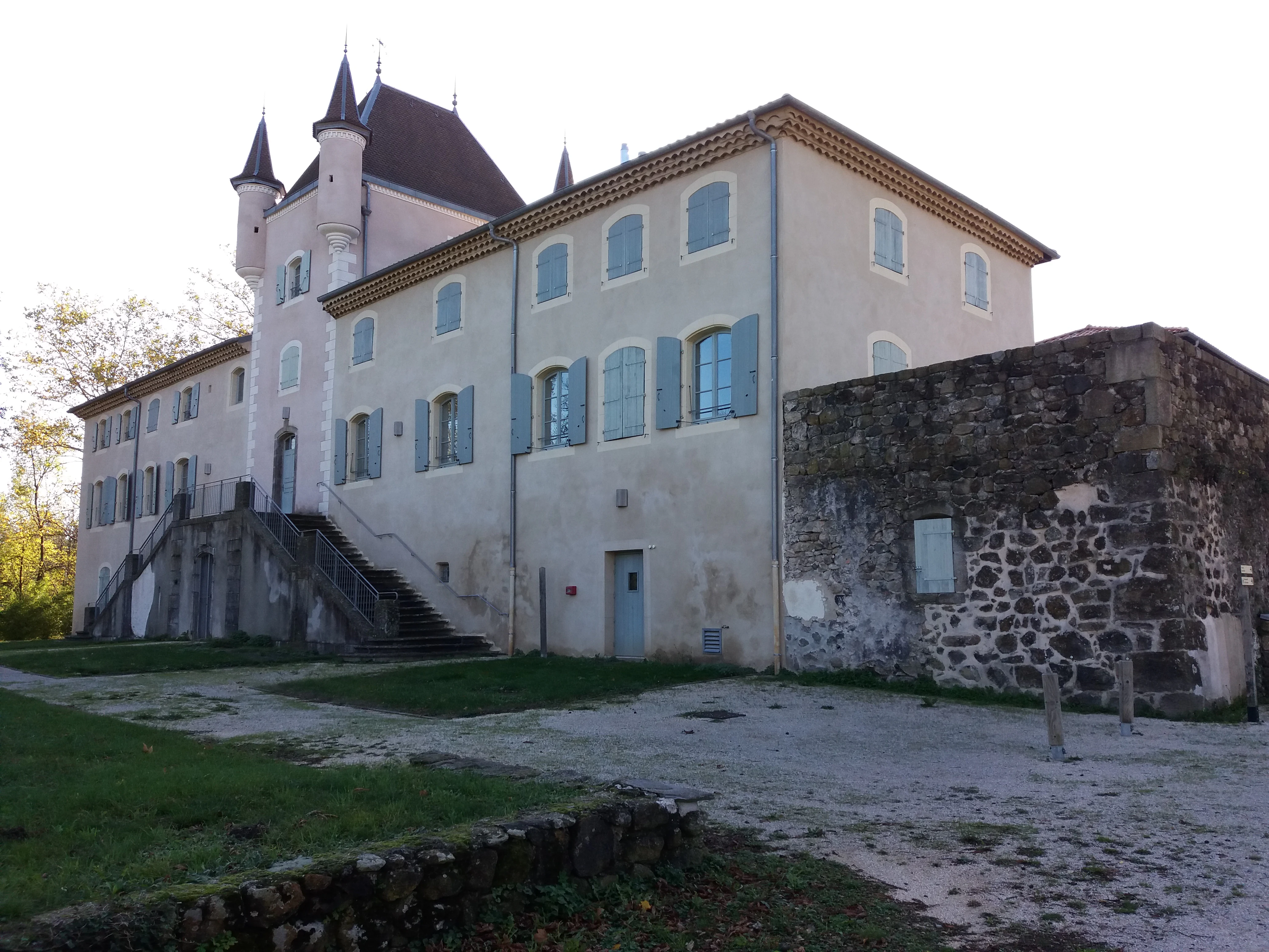 A multi-story historic chateau building with light-colored walls and blue shutters, featuring a stone tower section and an external stone staircase.