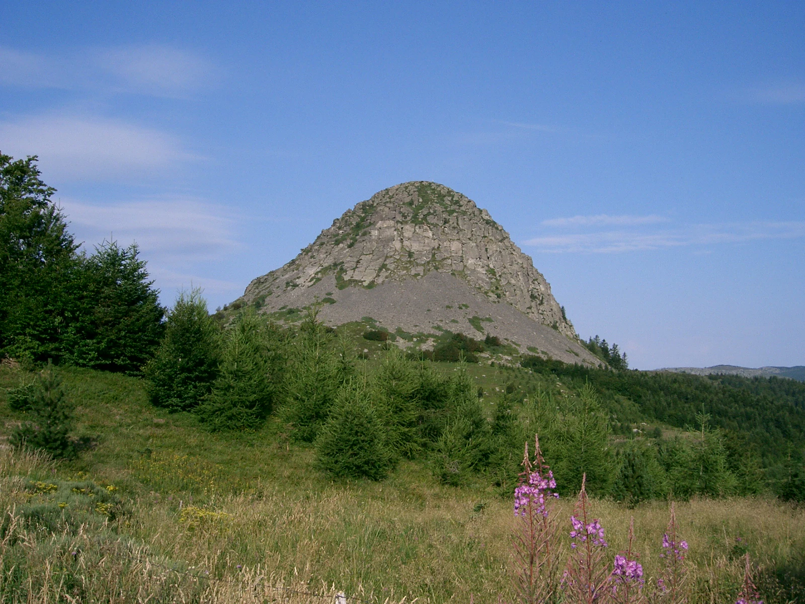 A wide angle view of the cone-shaped, rocky Mont Gerbier de Jonc mountain peak rising above a green, grassy, and forested landscape under a clear blue sky.