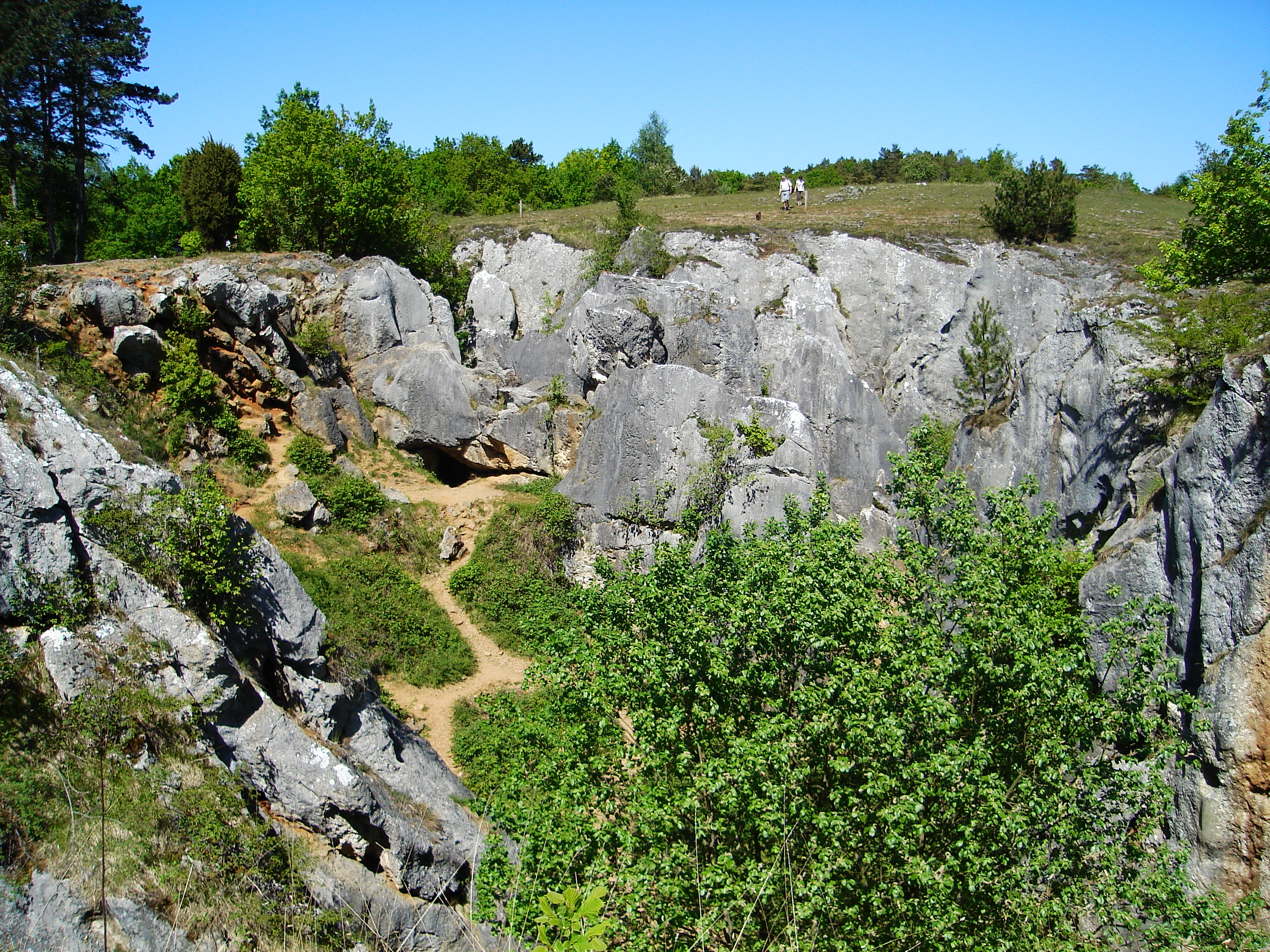 A sunny, eye-level landscape shot of a rocky limestone quarry or ravine with sparse vegetation, green trees at the edges, and a walking path winding through the rocks, with two small figures visible in the distance.