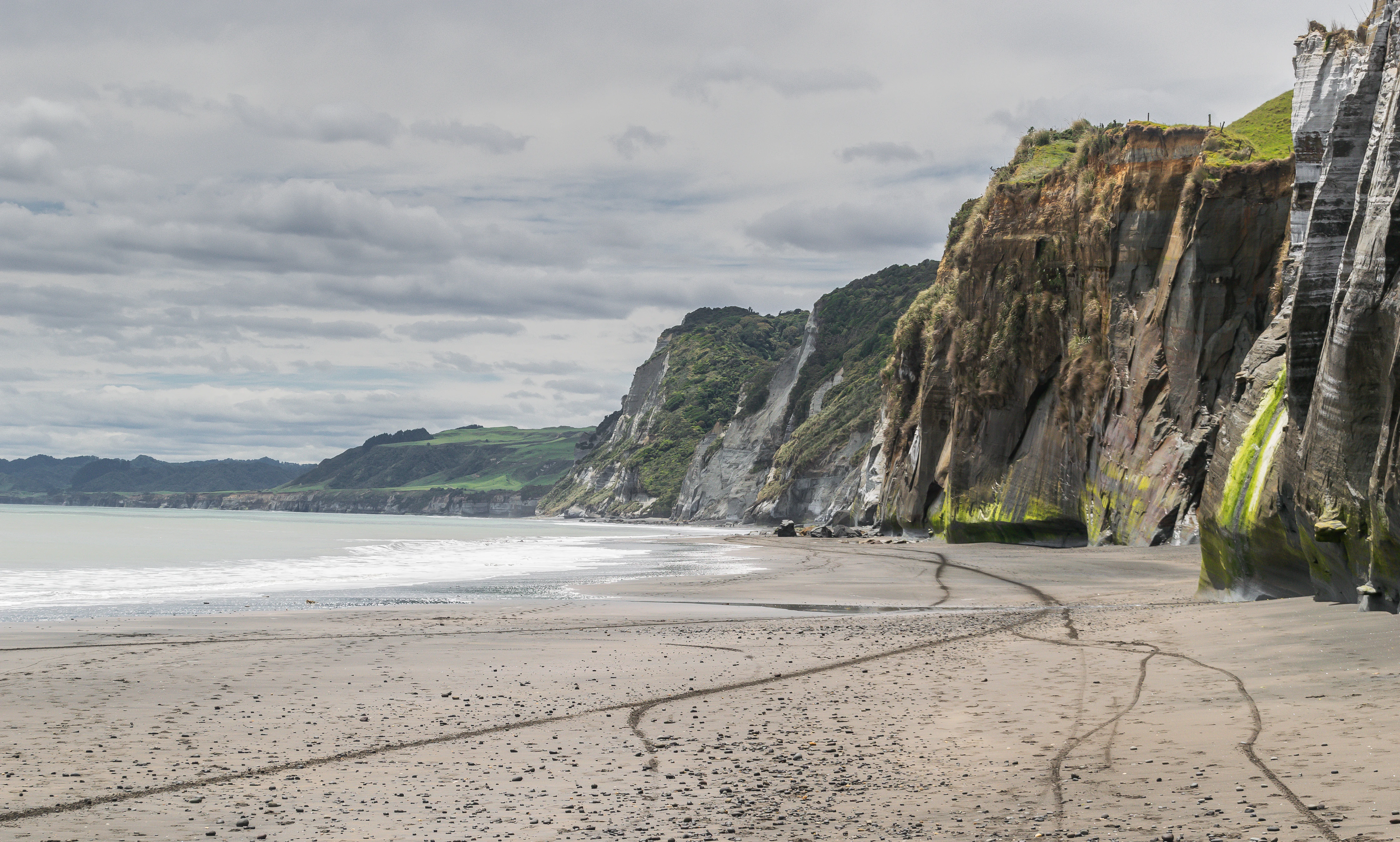 A wide shot of a sandy beach stretching toward towering, vertical coastal cliffs under a cloudy sky.