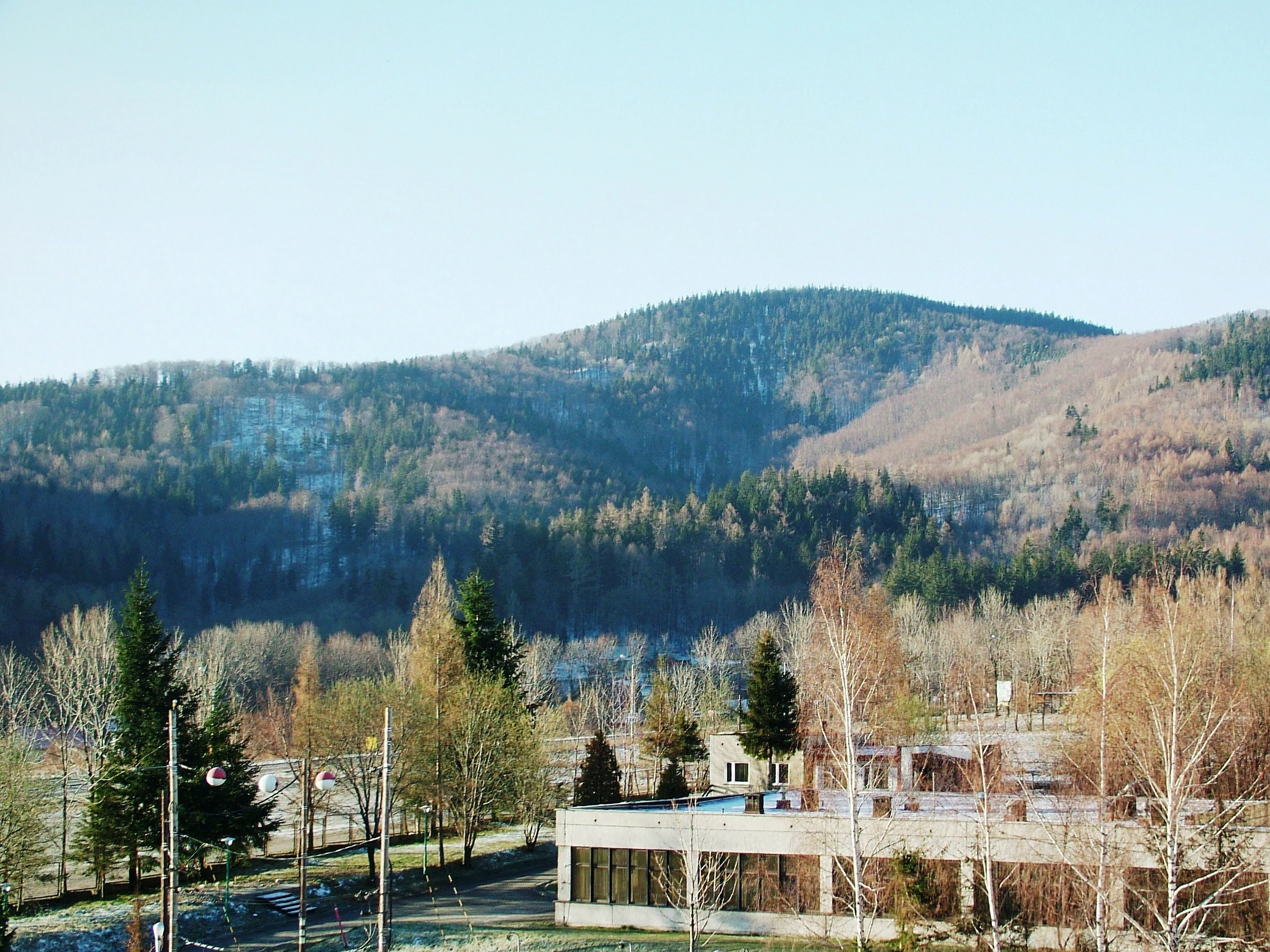 A wide view of a forested mountain slope known as Kolowrot peak in the Silesian Beskids, with a low-rise building and trees visible in the foreground.