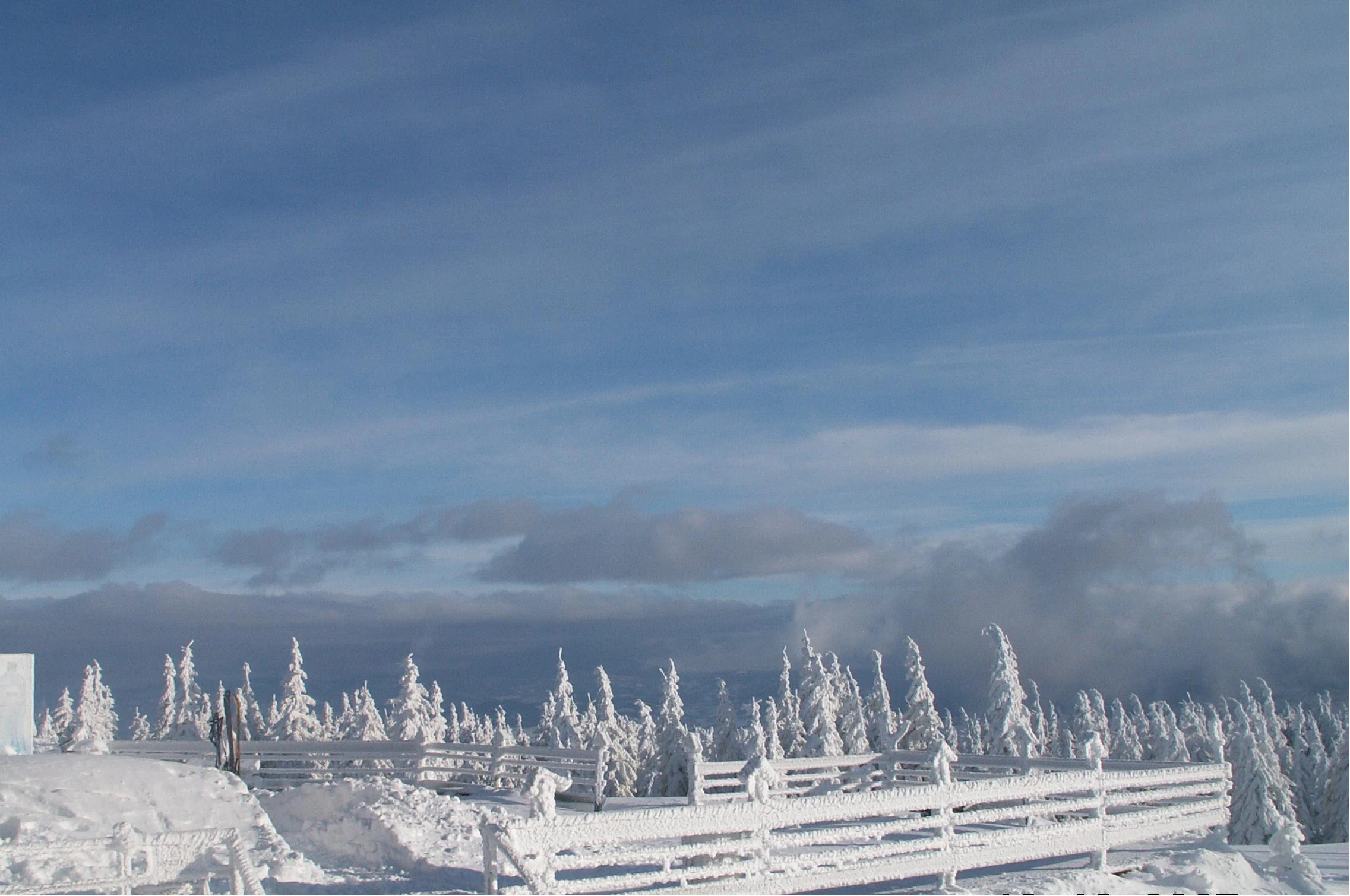 A wide shot of a snowy mountain summit with rime-covered trees, a wooden fence, and a blue sky with light clouds.