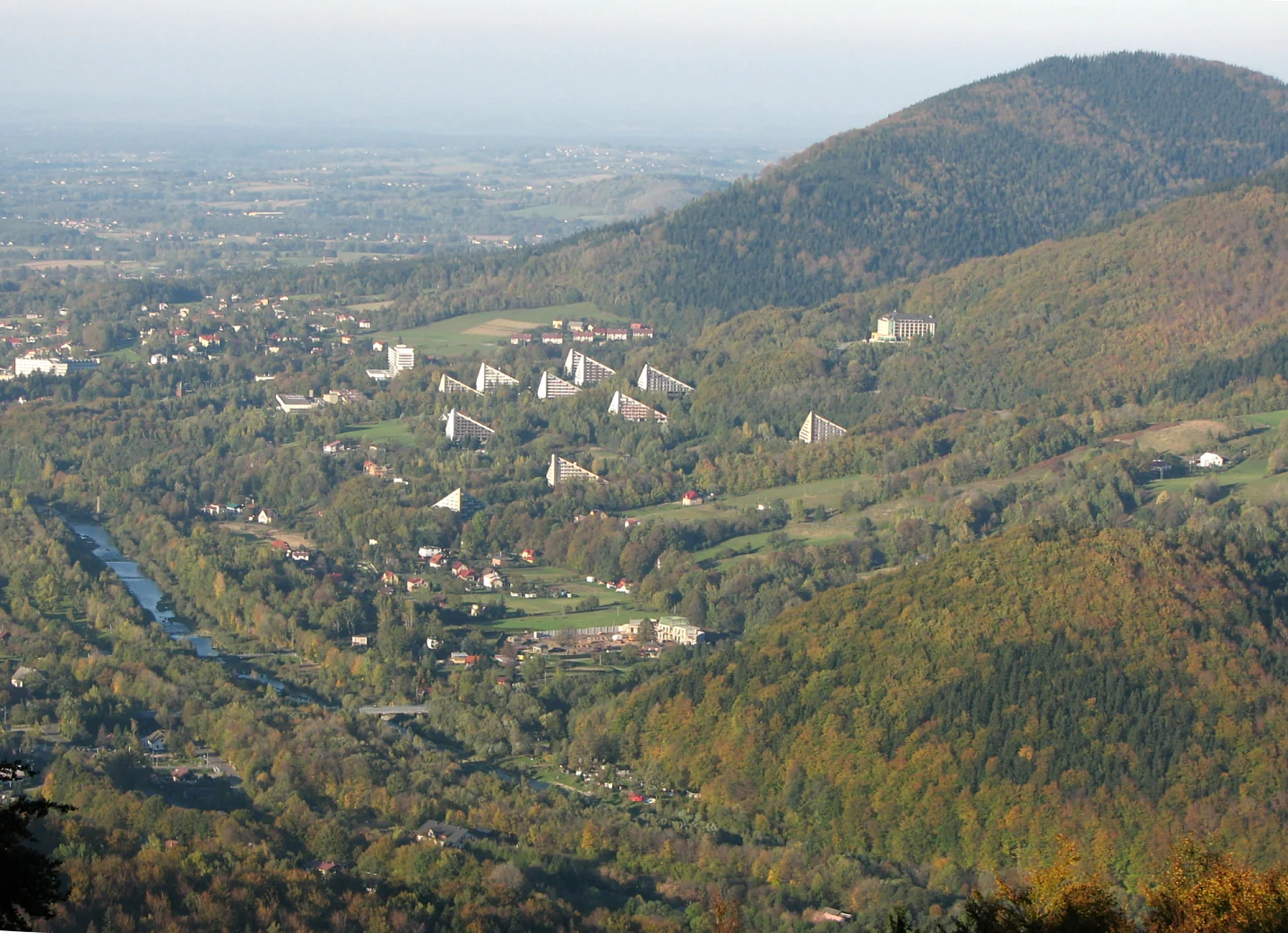 A wide angle high perspective view of a valley with forested mountains, a small town with several distinctive triangular buildings, and a river flowing through the center.