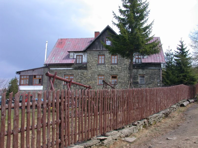A multi-story stone mountain hut with a red roof, enclosed by a rustic wooden picket fence, seen under a cloudy sky.