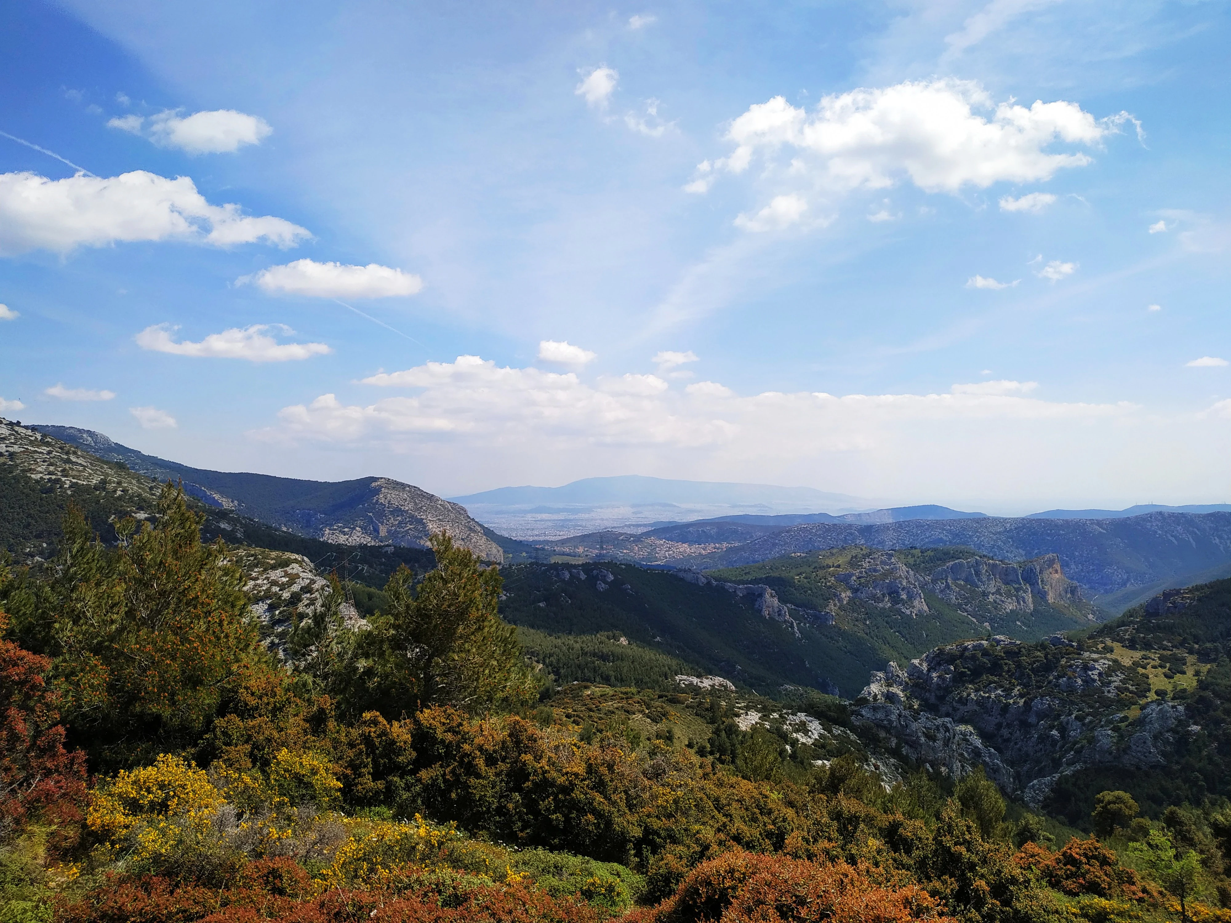 A wide, elevated view of mountainous terrain featuring forested slopes, rocky outcrops, and distant peaks under a blue sky with scattered clouds.
