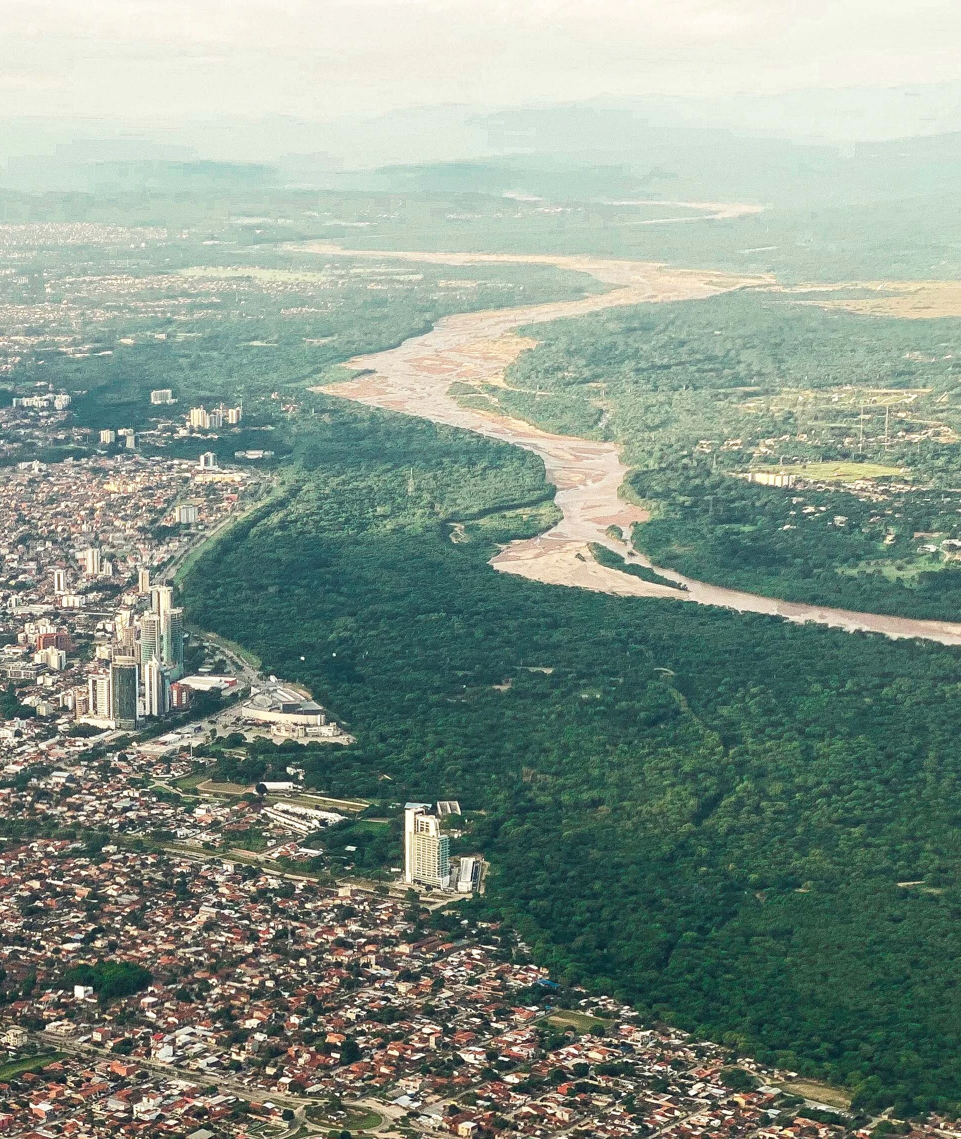Aerial view of the Piraí River winding through a vast green forest zone, adjacent to the densely built urban area of Santa Cruz de la Sierra.