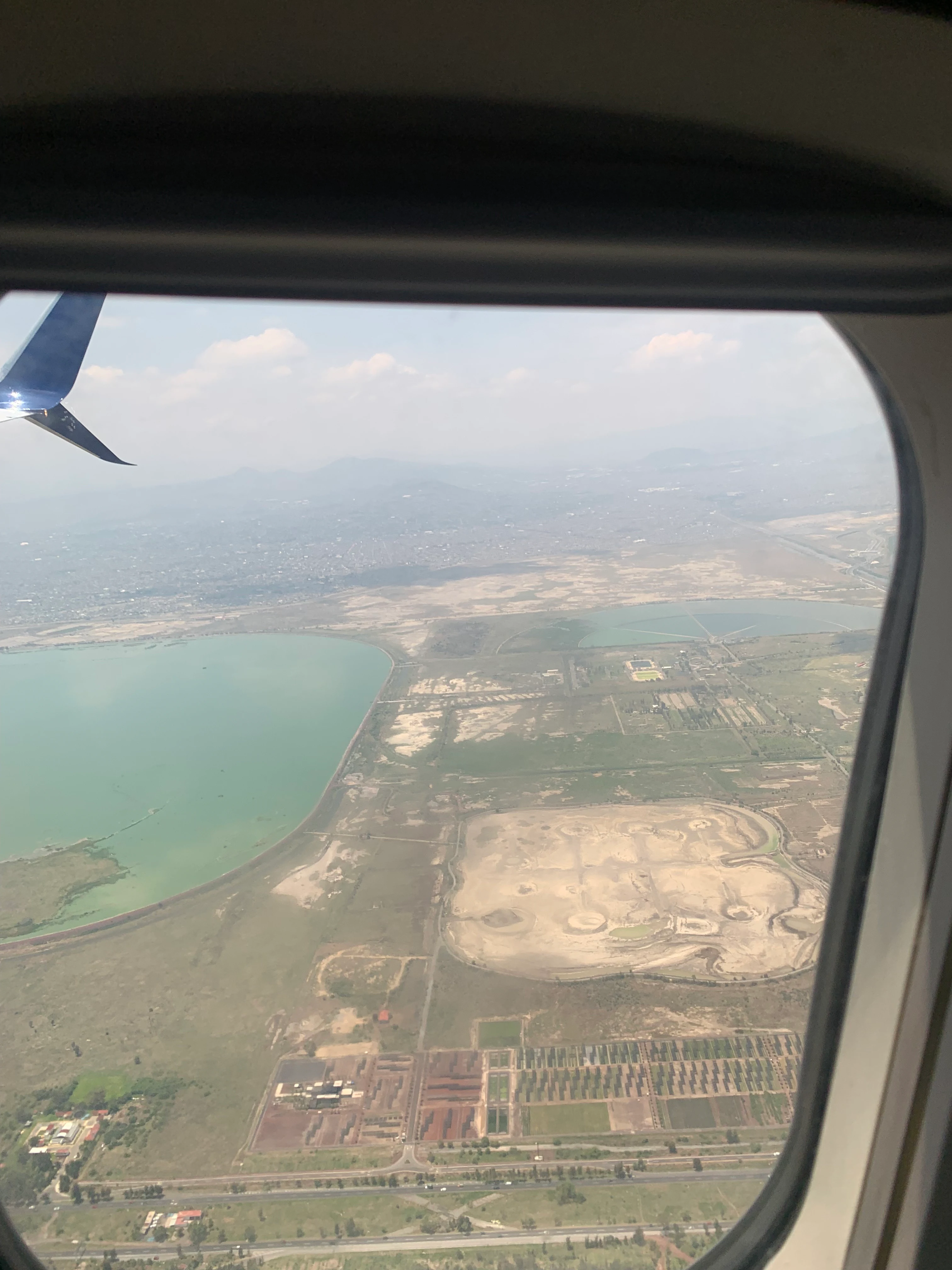 An aerial view of the Parque Ecológico Lago de Texcoco and the Nabor Carrillo reservoir, captured through an airplane window with a winglet visible in the upper left corner.
