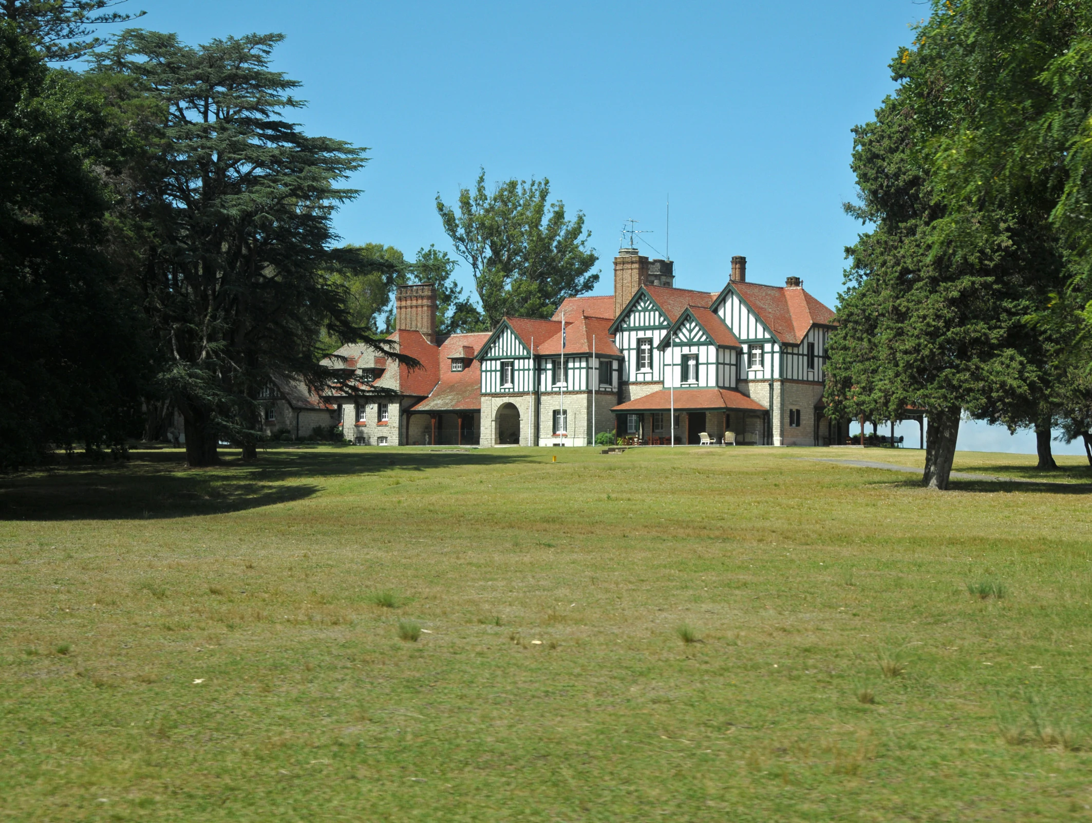 Una gran residencia de estilo Tudor de varias plantas, con estuco blanco y detalles de entramado de madera, situada en un amplio campo abierto y herboso rodeado de densos árboles maduros.