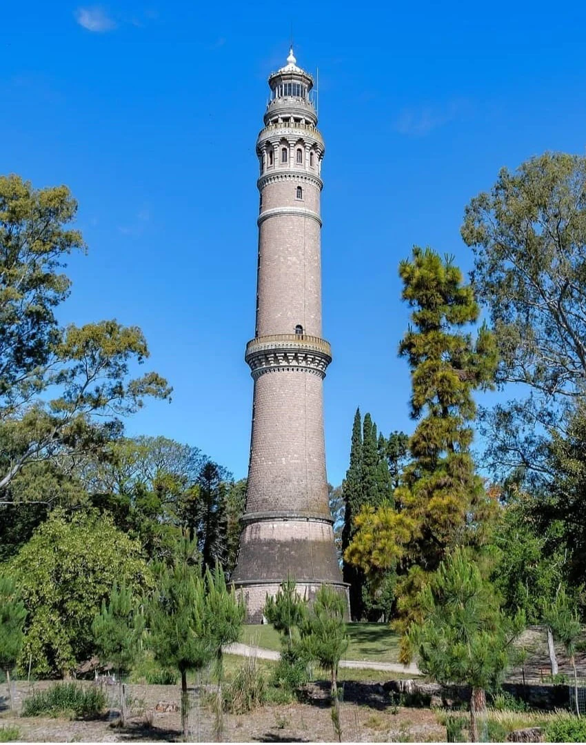 Una torre de ladrillo histórica y alta, la Torre Gaboto, se alza en el centro entre frondosos árboles verdes bajo un cielo azul despejado.