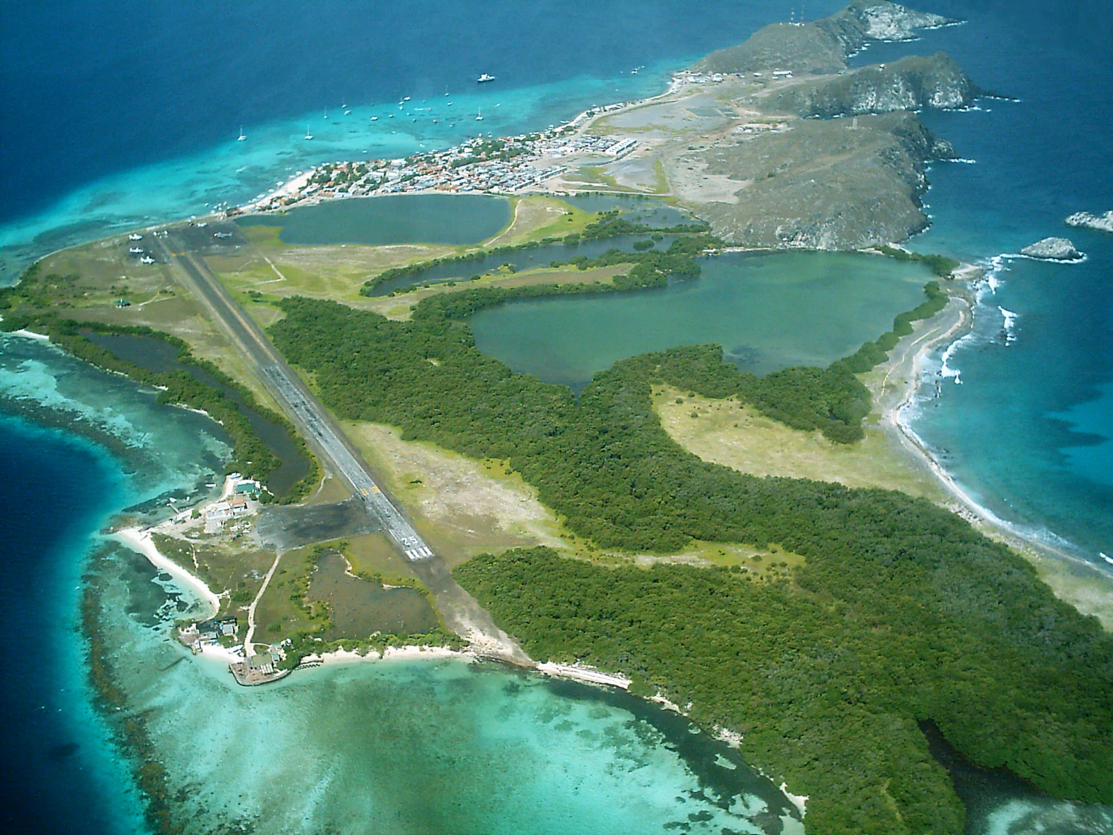 An aerial photograph of a tropical island featuring a long airstrip, green vegetation, several inland lagoons, and turquoise shallow waters surrounding the coastline.