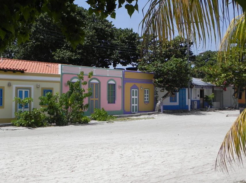 A wide white sandy street lined with colorful single-story buildings painted in yellow, pink, blue, and orange, framed by palm fronds and tropical trees under a clear blue sky.