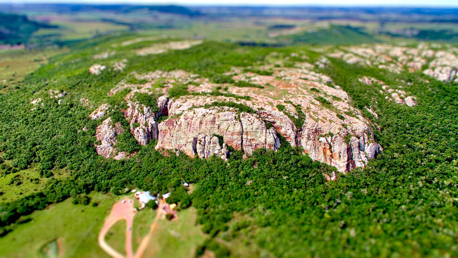 A high-angle aerial view of a large, craggy volcanic rock formation covered in patches of forest, rising above green fields in a rural landscape.