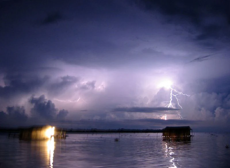 A night landscape view showing a bright lightning bolt striking the horizon above a calm dark body of water with stilt houses silhouetted in the distance.