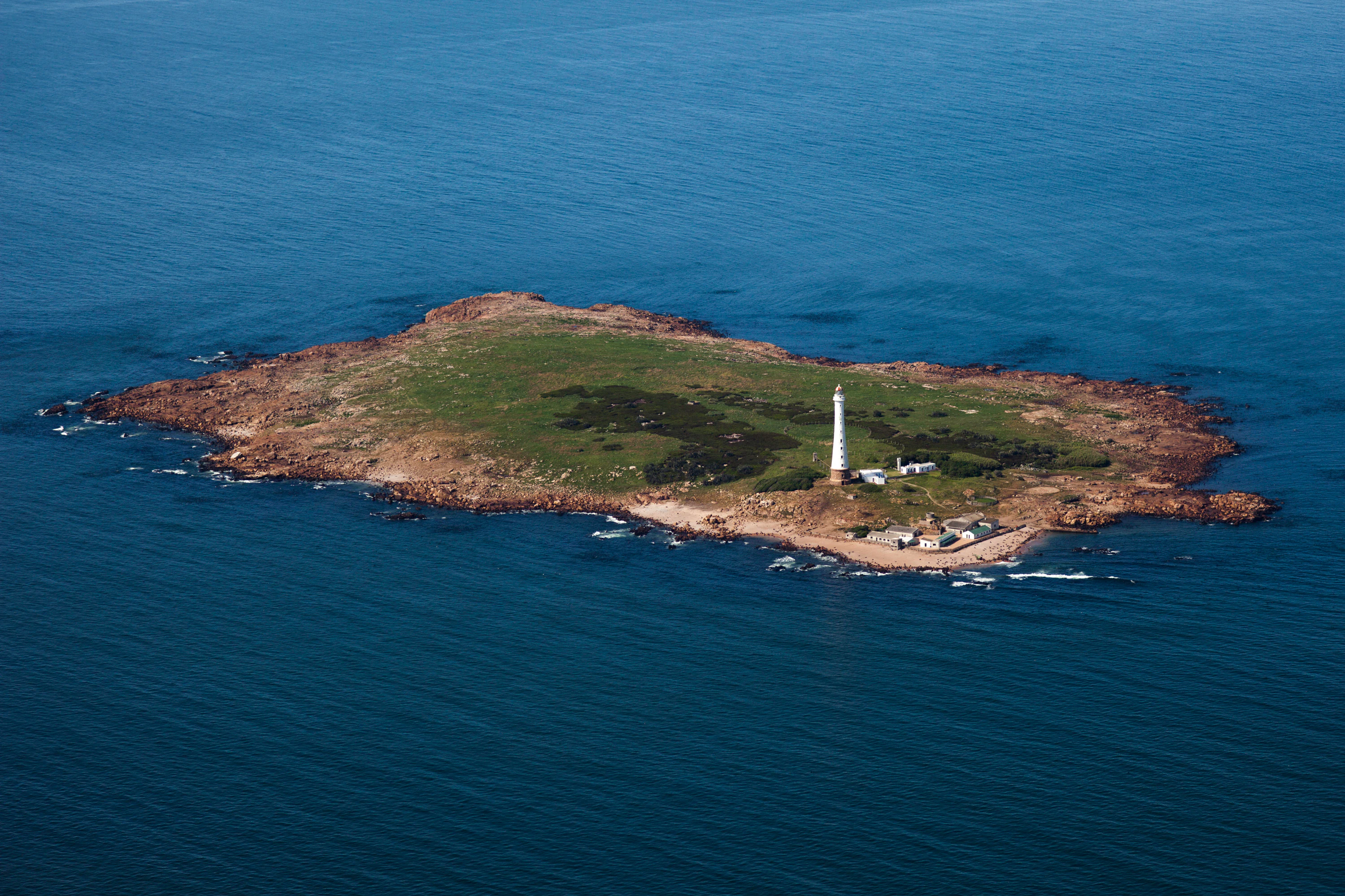 Una vista aérea de la rocosa Isla de Lobos rodeada de aguas oceánicas de un azul profundo, con un alto faro blanco visible en la superficie de la isla.