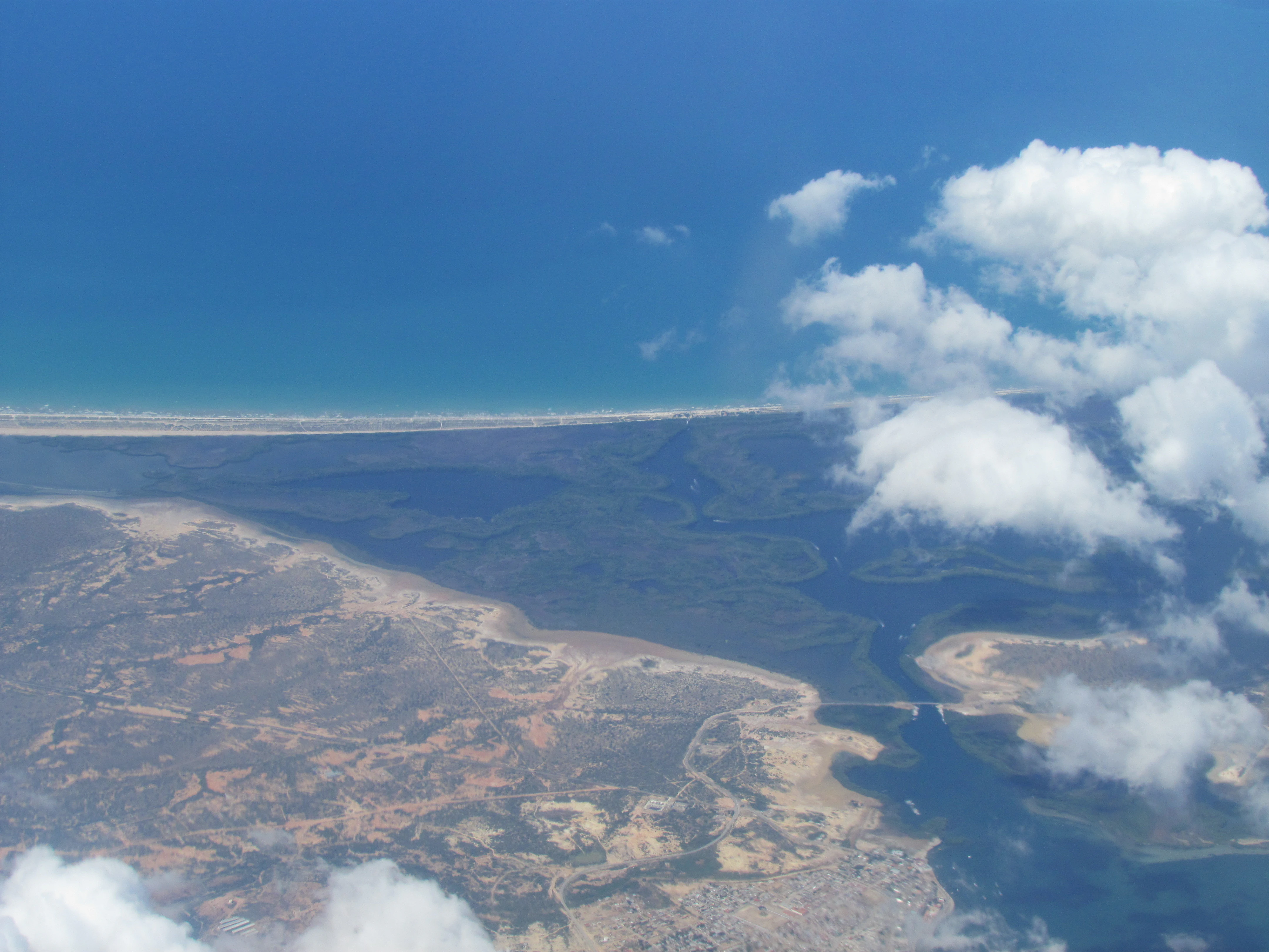 Aerial view of a coastal lagoon with a narrow sandy barrier beach separating it from the blue ocean, surrounded by dark mangrove vegetation.