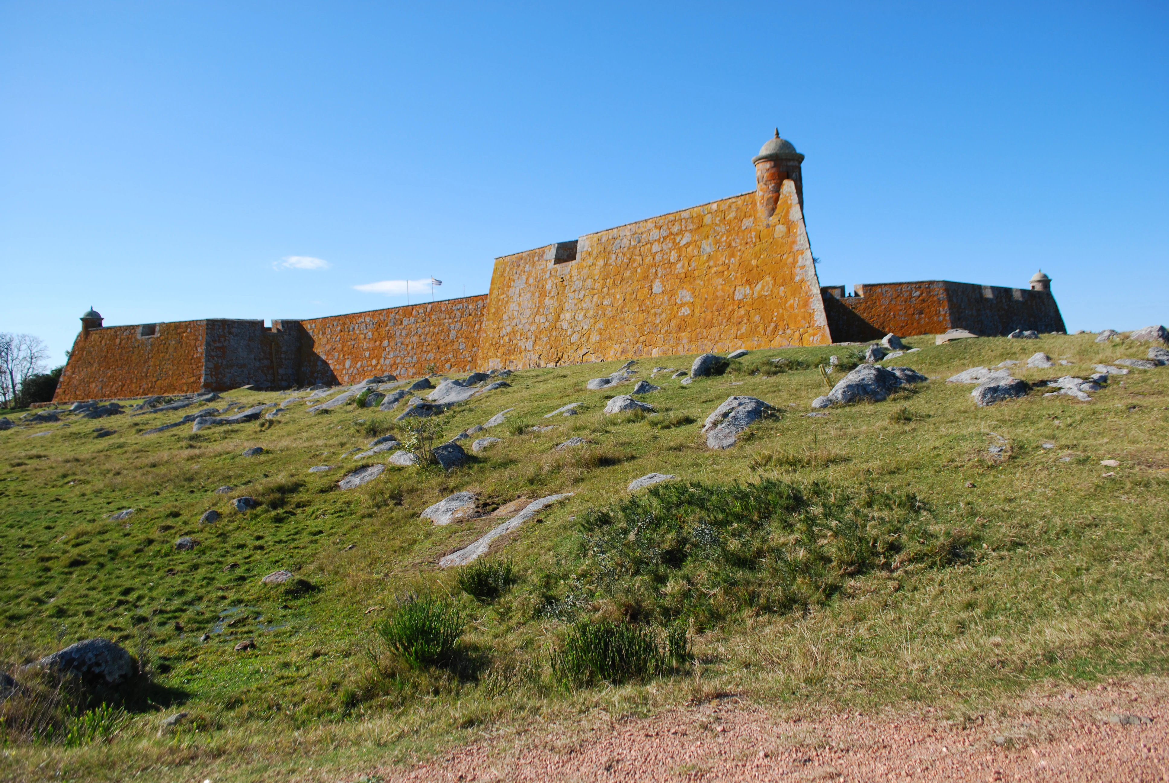 A historic fort with orange-colored stone walls and a rounded watchtower sitting atop a grassy hill scattered with rocks, under a clear blue sky.