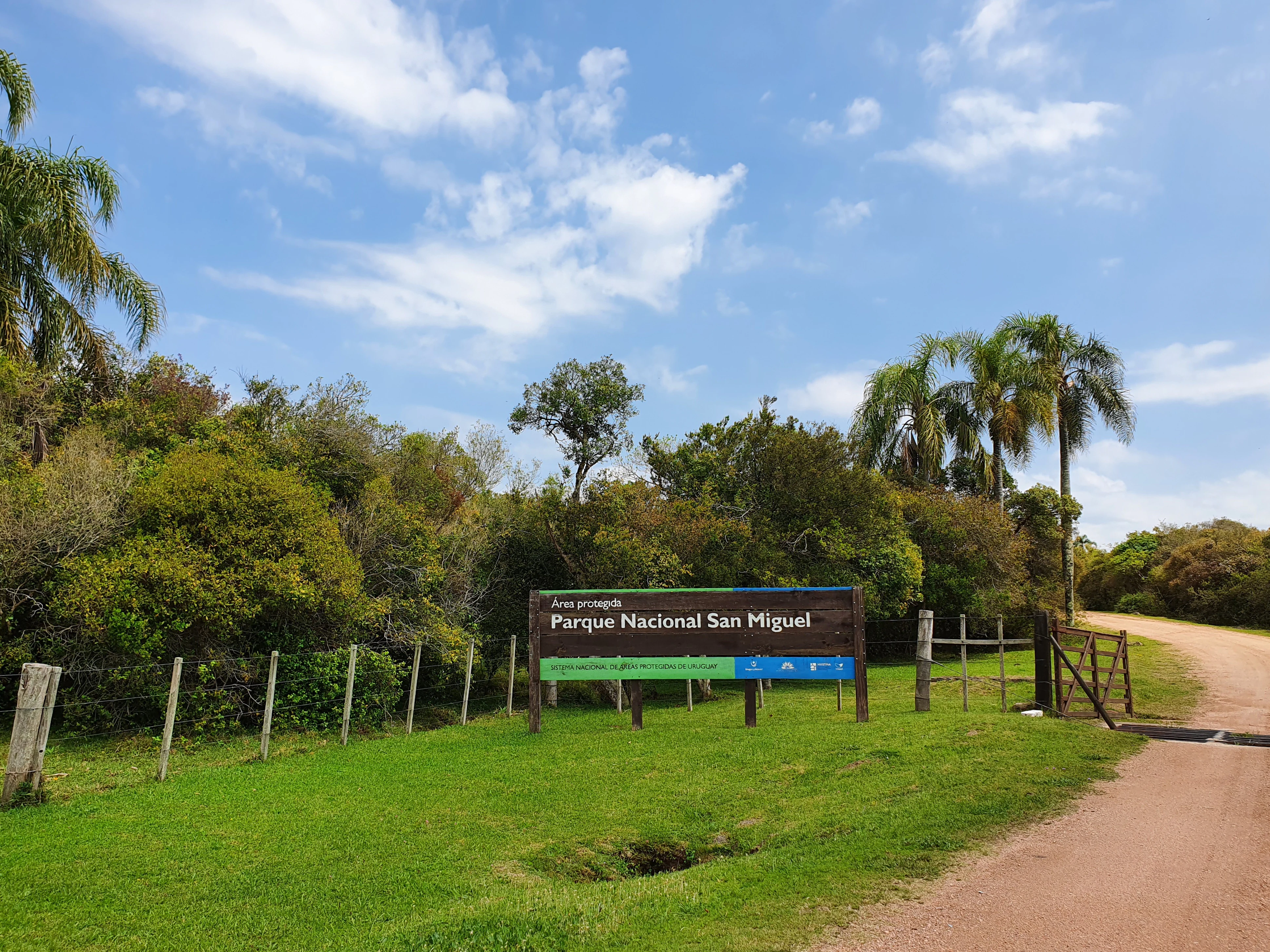 A wooden sign reading Parque Nacional San Miguel stands beside a dirt road entrance, surrounded by green grass, trees, and a blue sky with clouds.