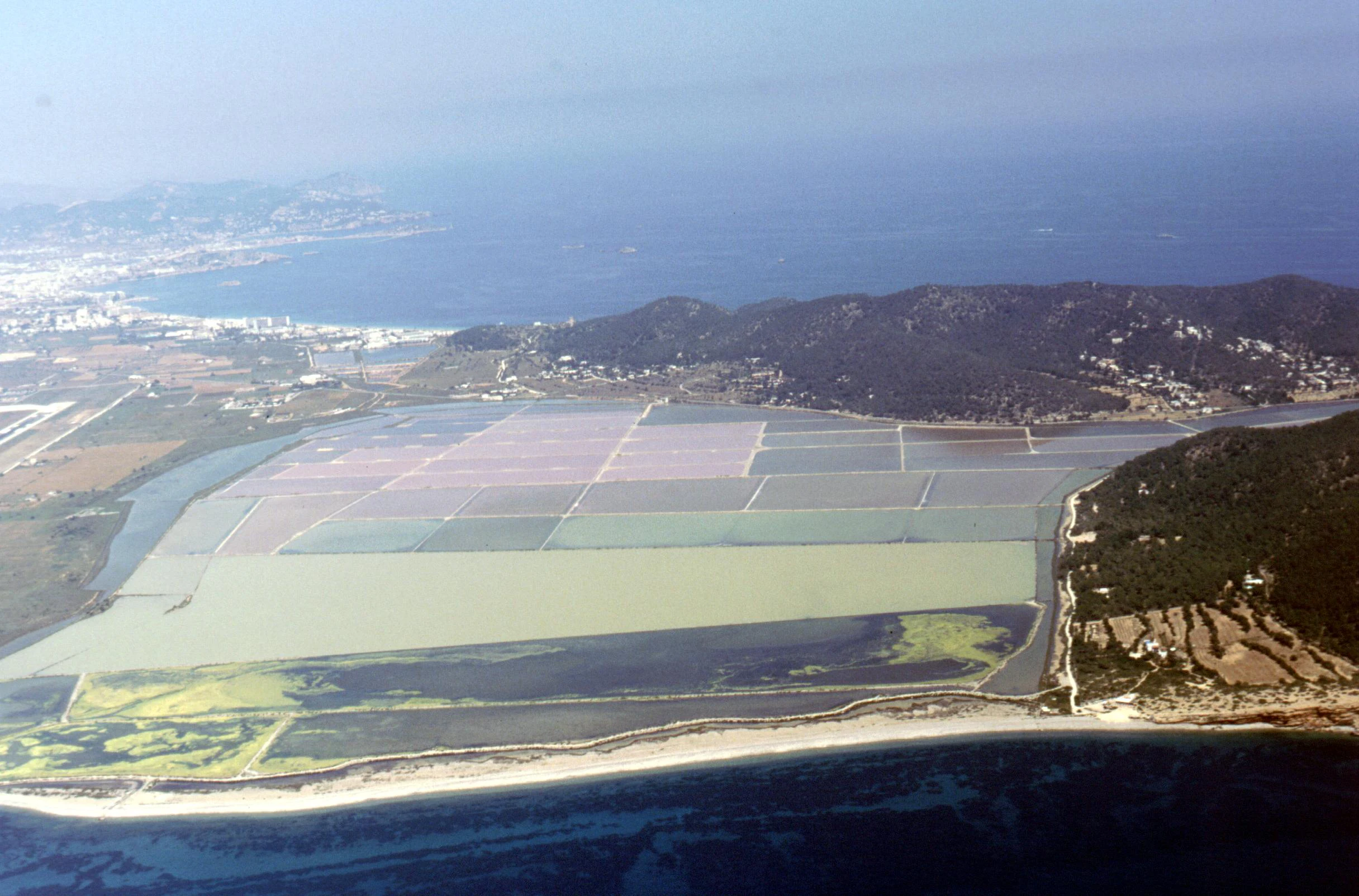 An aerial view showing rectangular salt evaporation ponds divided by levees, next to a coastline and hilly forested terrain.