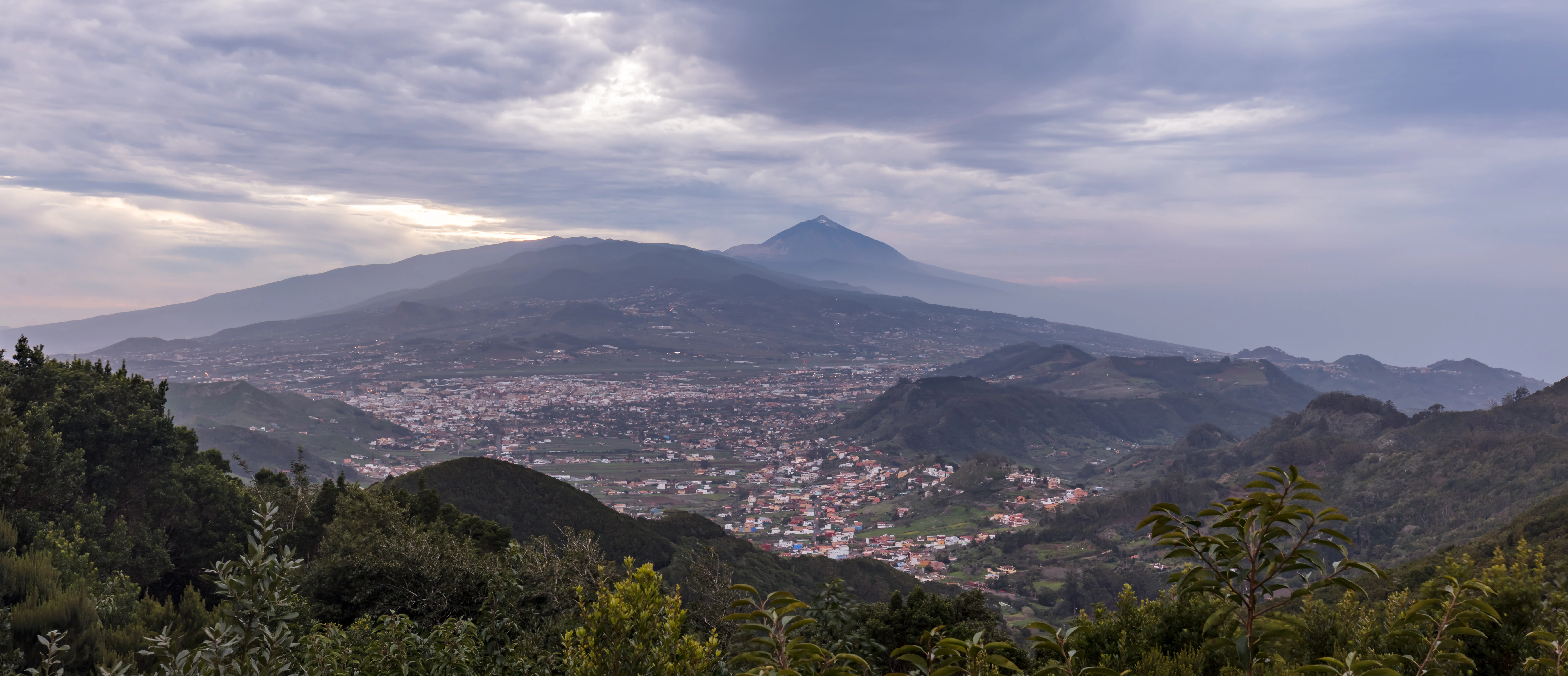 A wide landscape view of the Anaga massif featuring dense green forested ridges in the foreground and a town settled in the valley below, with the prominent peak of Mount Teide visible in the hazy, cloudy background.