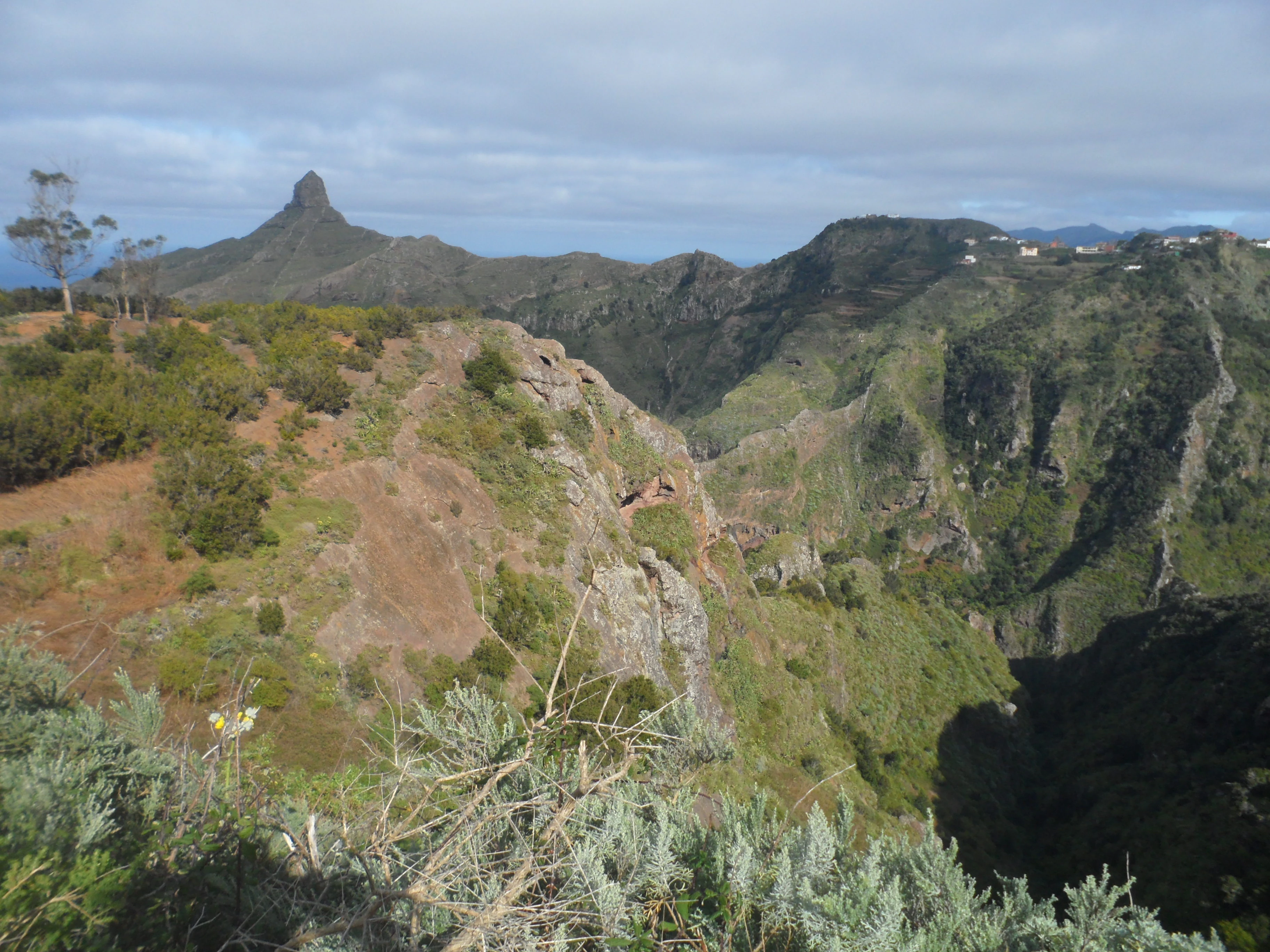 A wide, panoramic landscape view of a green mountainous region with a prominent, sharp, cone-shaped rock peak in the background and a valley in the foreground.