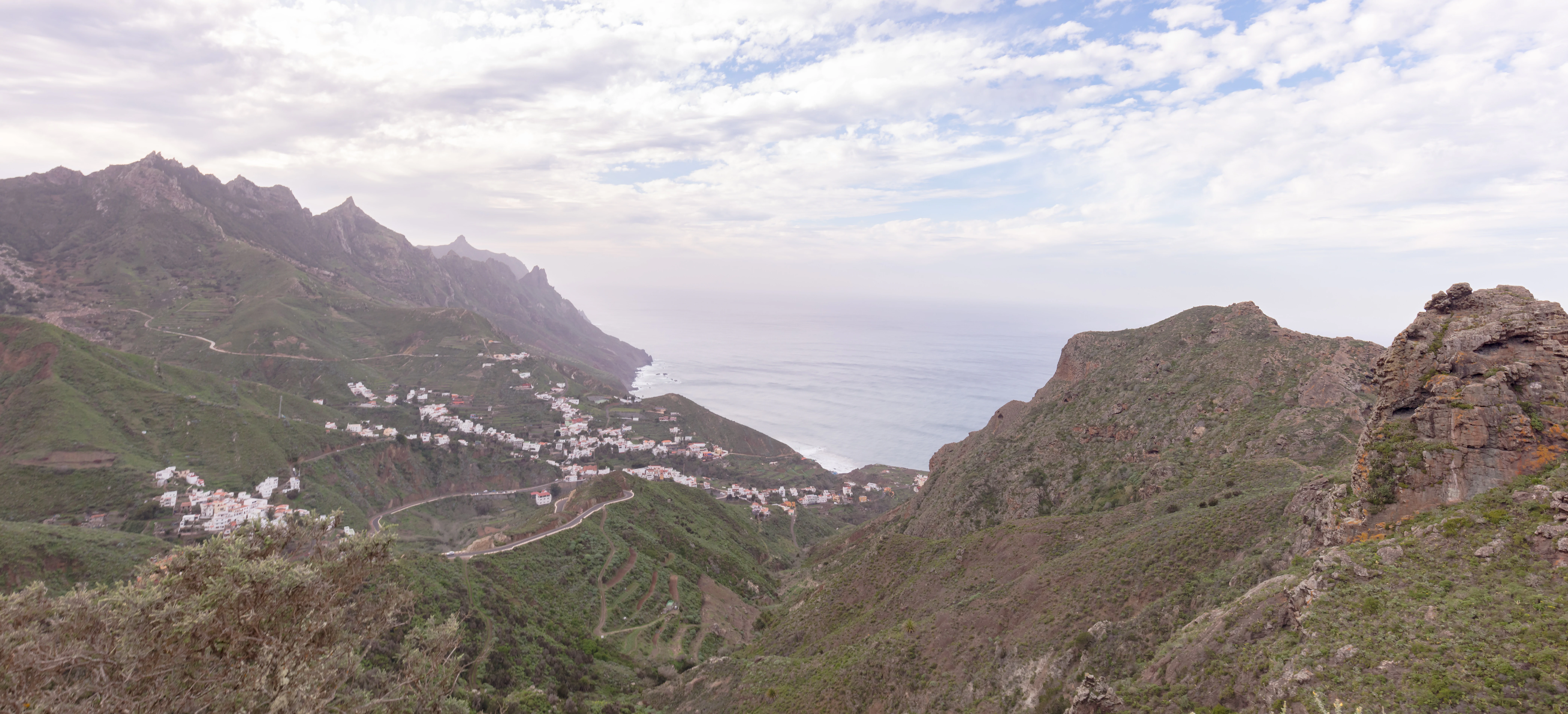 Wide landscape view of mountainous terrain with green slopes, a small village of white buildings nestled on a hillside, and the ocean visible in the distance under a cloudy sky.