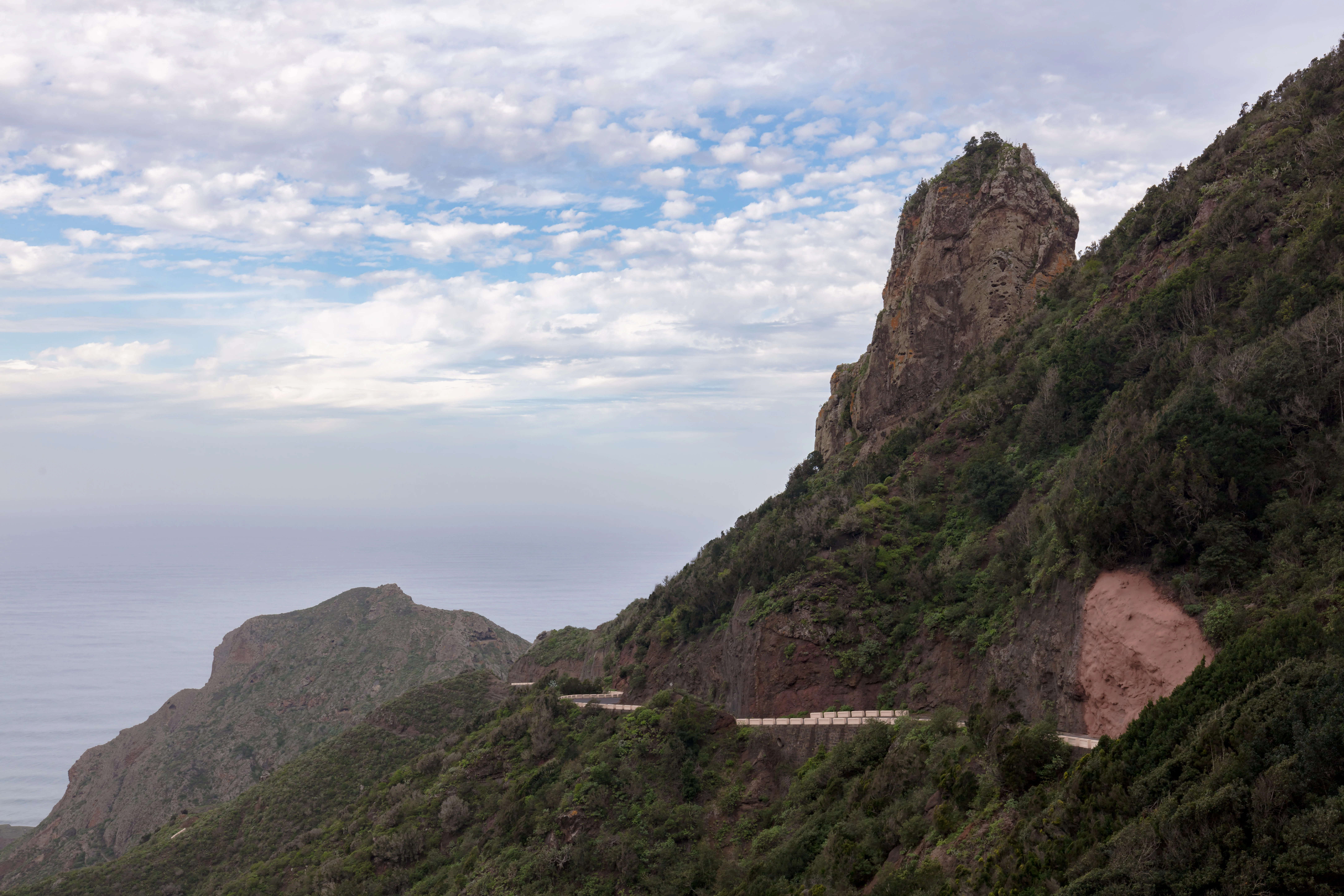 A steep, rugged mountain slope with a winding road built into its side, overlooking a hazy coastal horizon under a cloudy sky.
