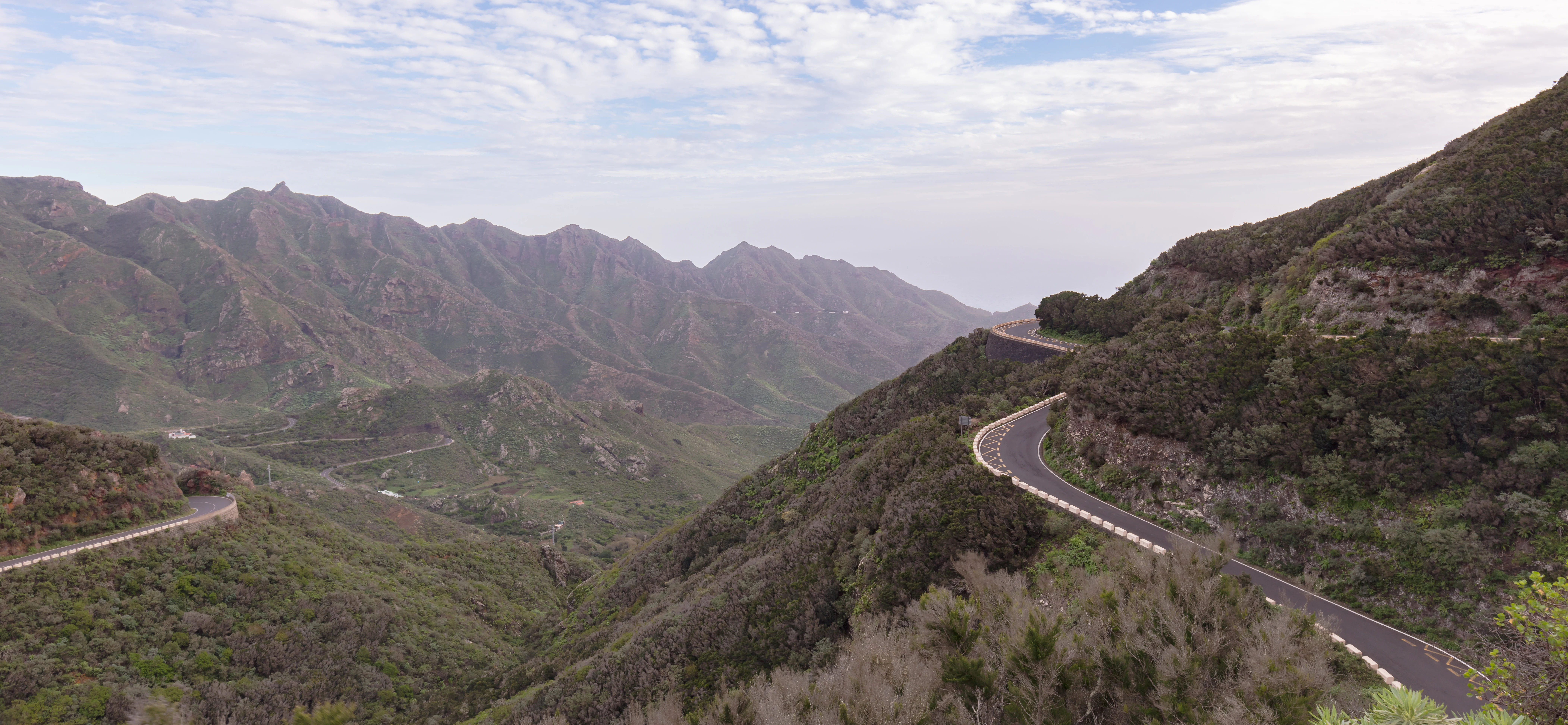 A paved mountain road winds through a steep, green valley with craggy ridgelines and forest vegetation under a cloudy sky.