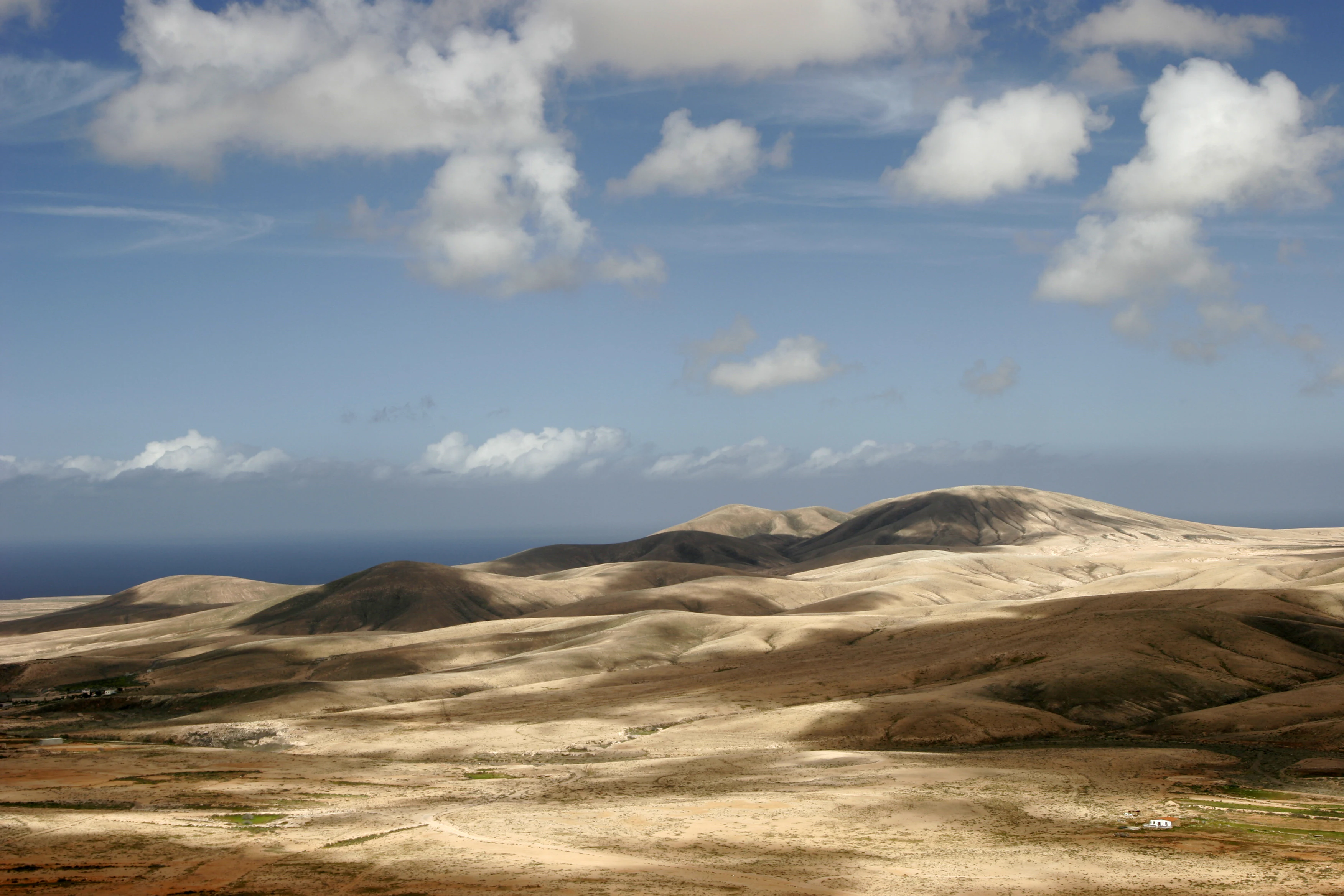 A wide, eye-level photograph of a dry, rolling landscape with brownish hills under a blue sky with scattered white clouds, showing the sea in the distant background.