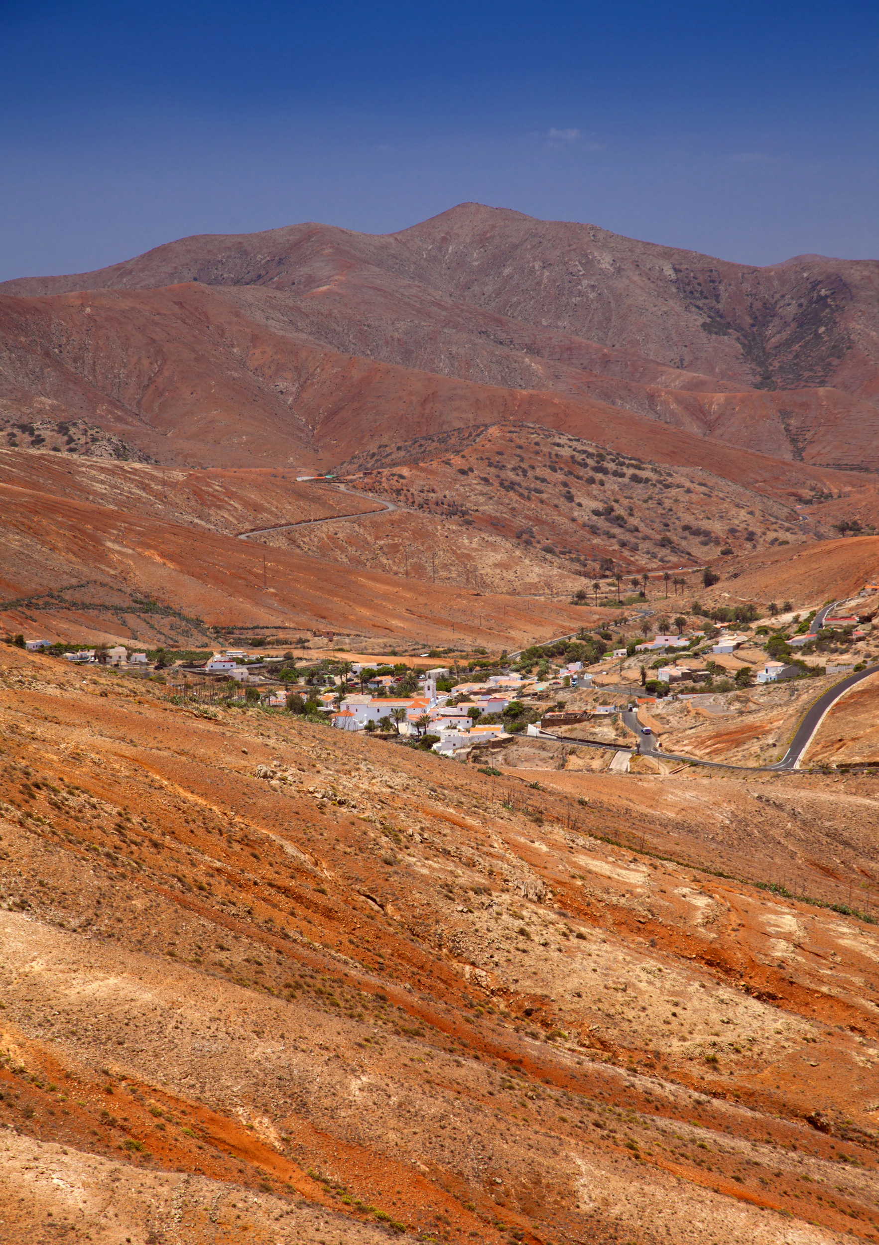 A wide, high-angle view showing a small village with white-walled buildings set in a deep valley surrounded by barren, reddish-brown volcanic mountain slopes under a clear blue sky.
