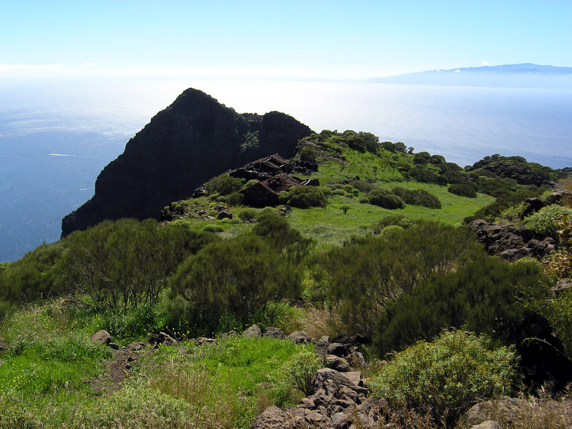 A high angle view of a grassy, volcanic ridgeline with small stone ruins, overlooking the Atlantic Ocean with a distant island visible on the horizon.