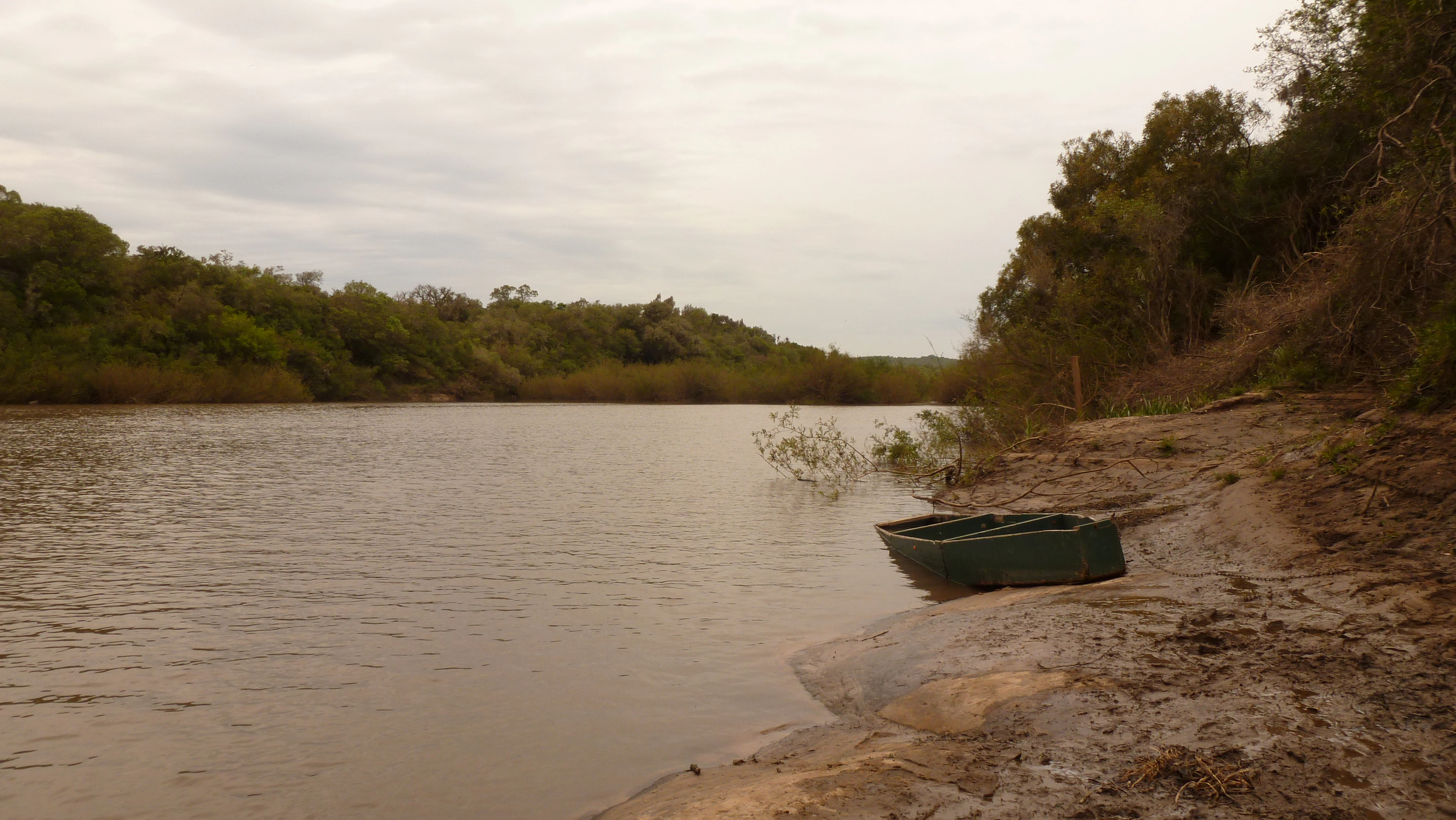 A small, dark boat sits at the edge of the Yaguaron River with a forested riverbank in the background under an overcast sky.