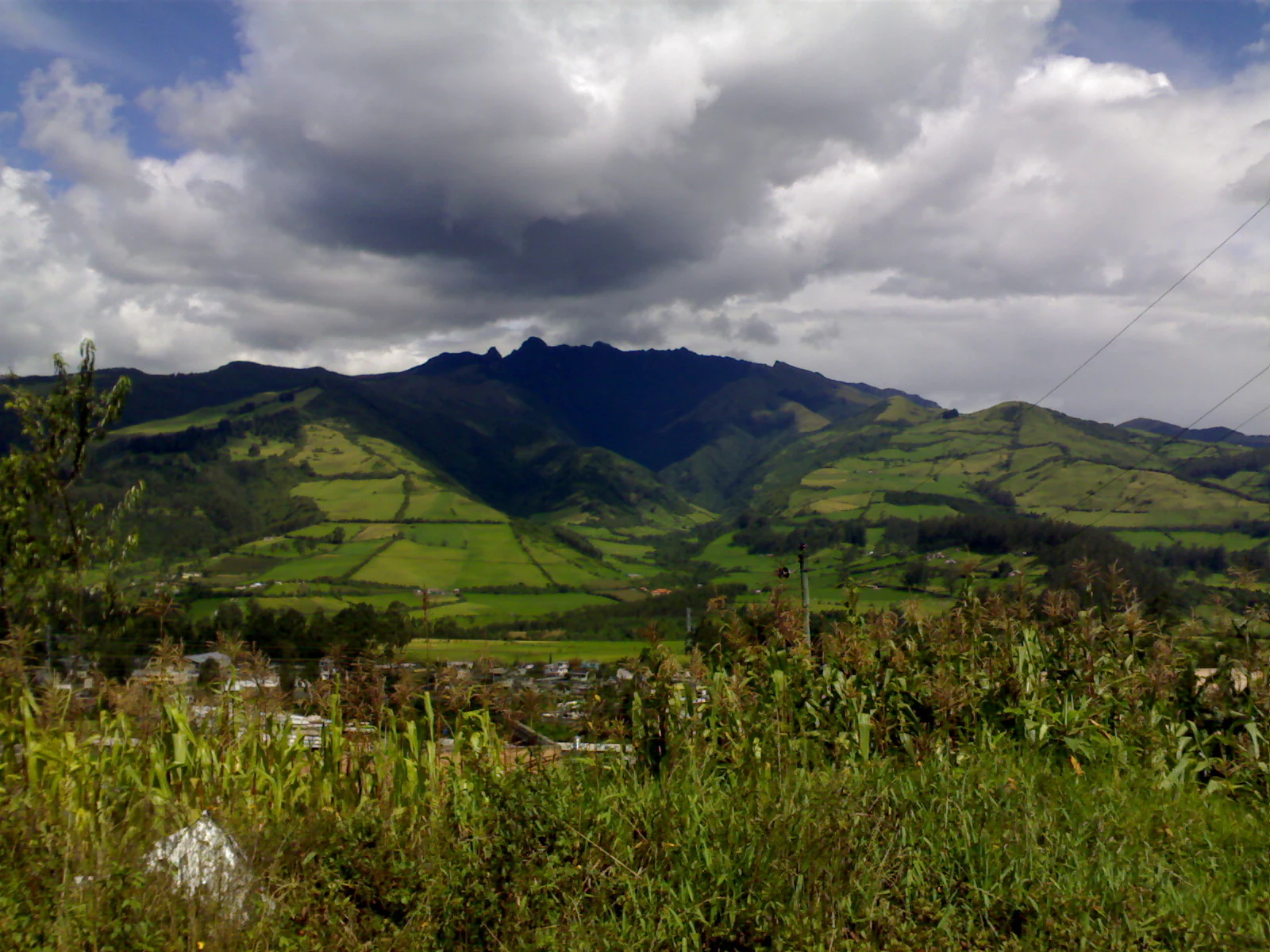 A wide shot of the Pasochoa volcano, a rugged, dark-forested mountain ridge rising above green fields and agricultural land under a cloudy sky.