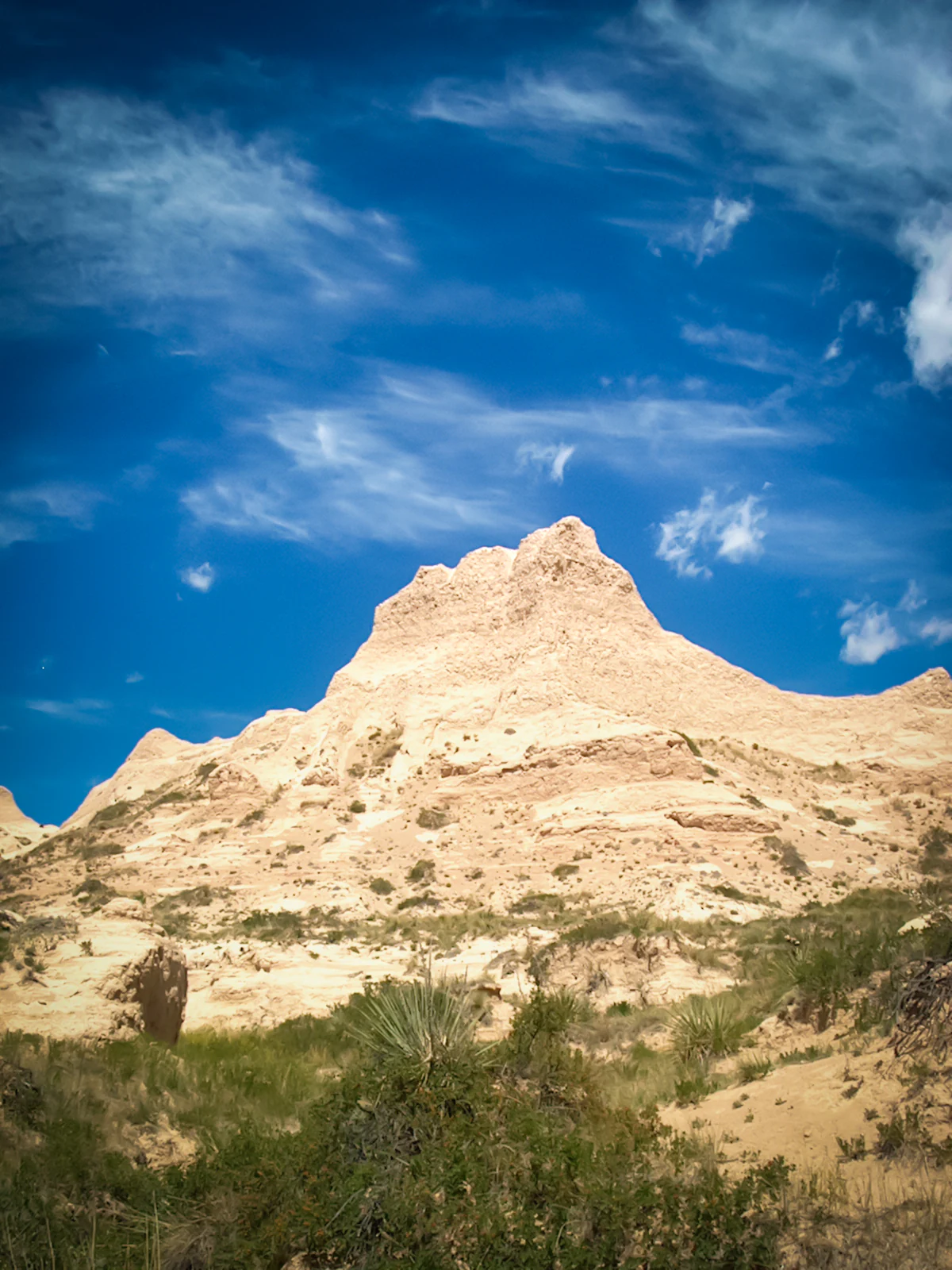 A steep, light-colored sedimentary butte stands against a vibrant blue sky with scattered clouds, surrounded by sparse prairie vegetation.