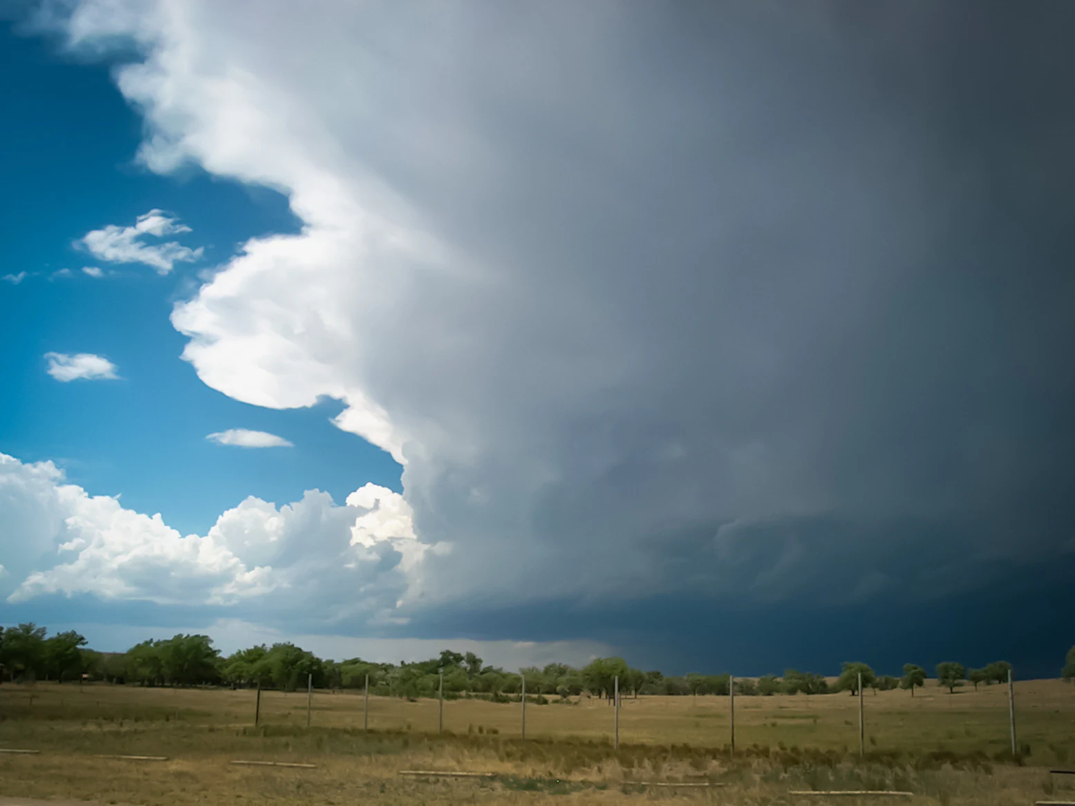 A large dark storm cloud looms over a flat, grassy plain with a simple wire fence in the foreground.