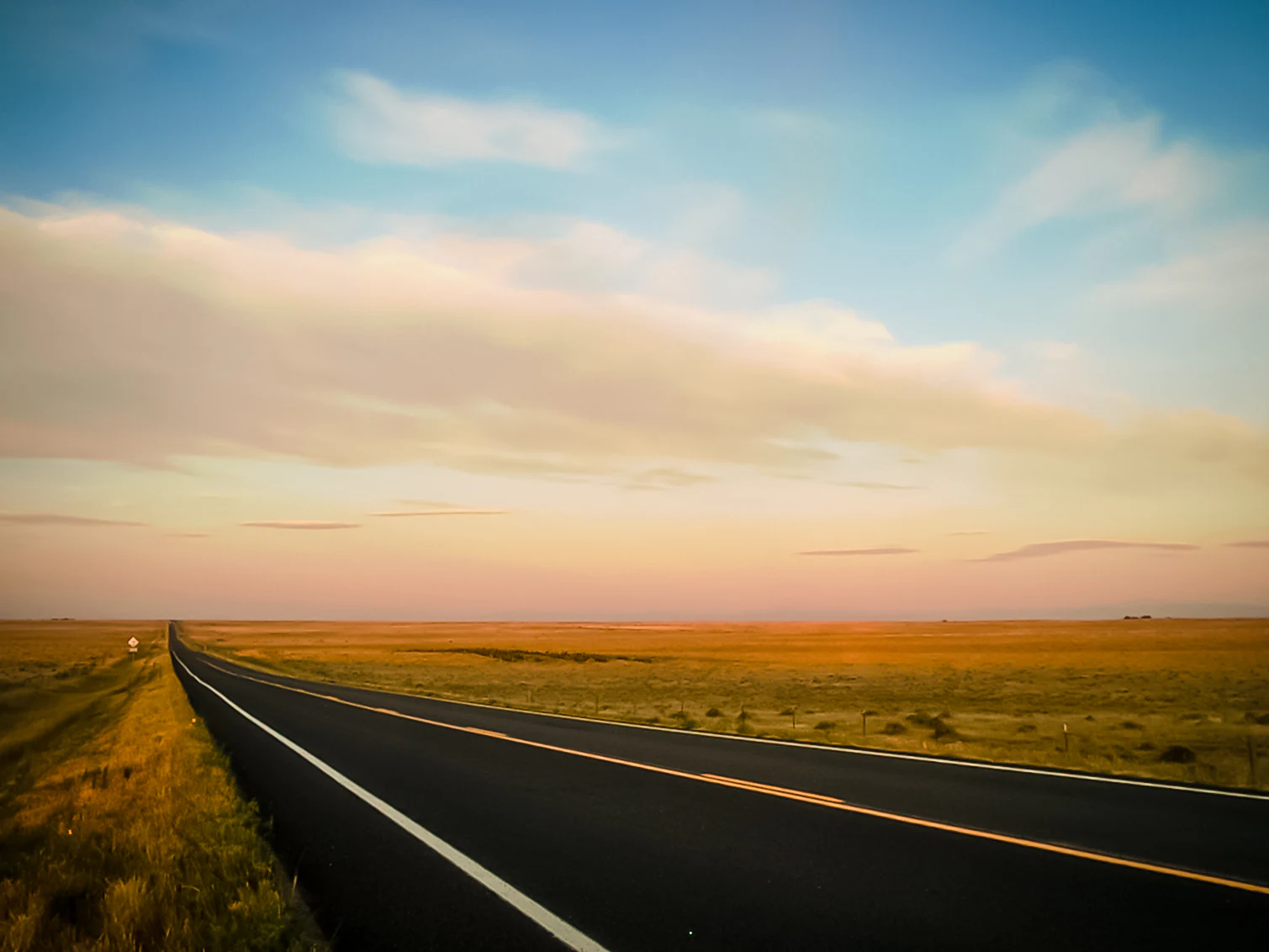 A long, straight paved road cuts through a vast, flat grassland under a soft sunrise sky with pink and blue hues.
