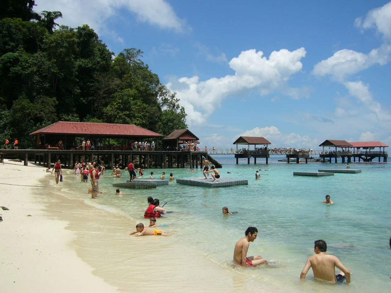 A group of people swimming and relaxing on a white sandy beach with wooden jetties and huts extending over the turquoise water.