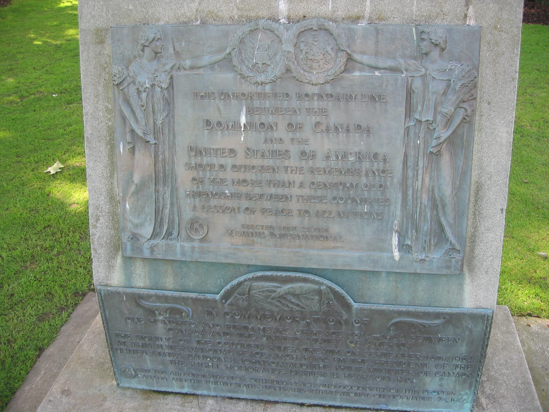 A bronze historical plaque set into a concrete base at Peace Arch Park, featuring embossed text and figures representing the peaceful relationship between Canada and the United States.