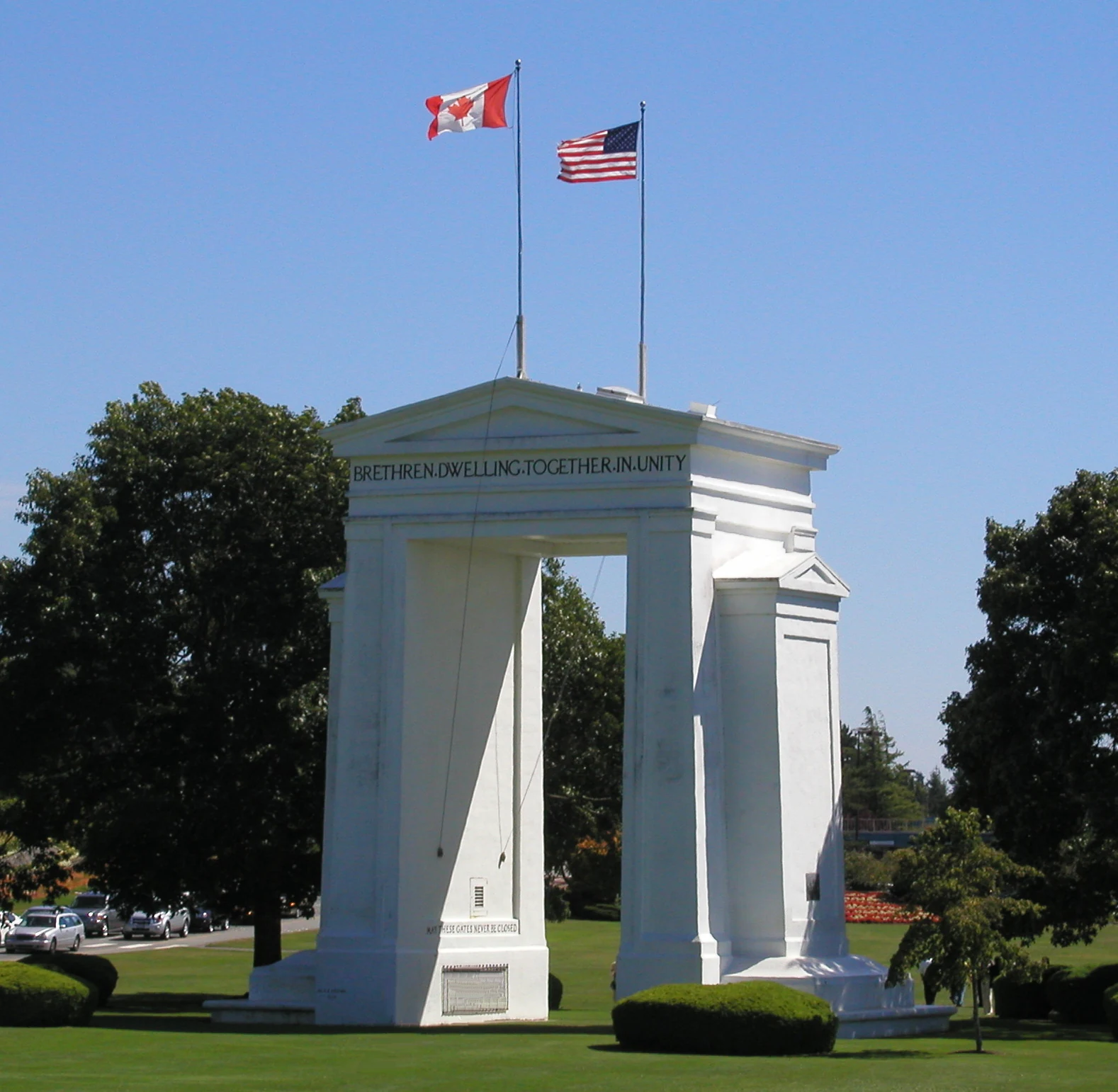 A white neoclassical monument titled Peace Arch stands in a grassy park, with the flags of Canada and the United States flying above it against a clear blue sky.