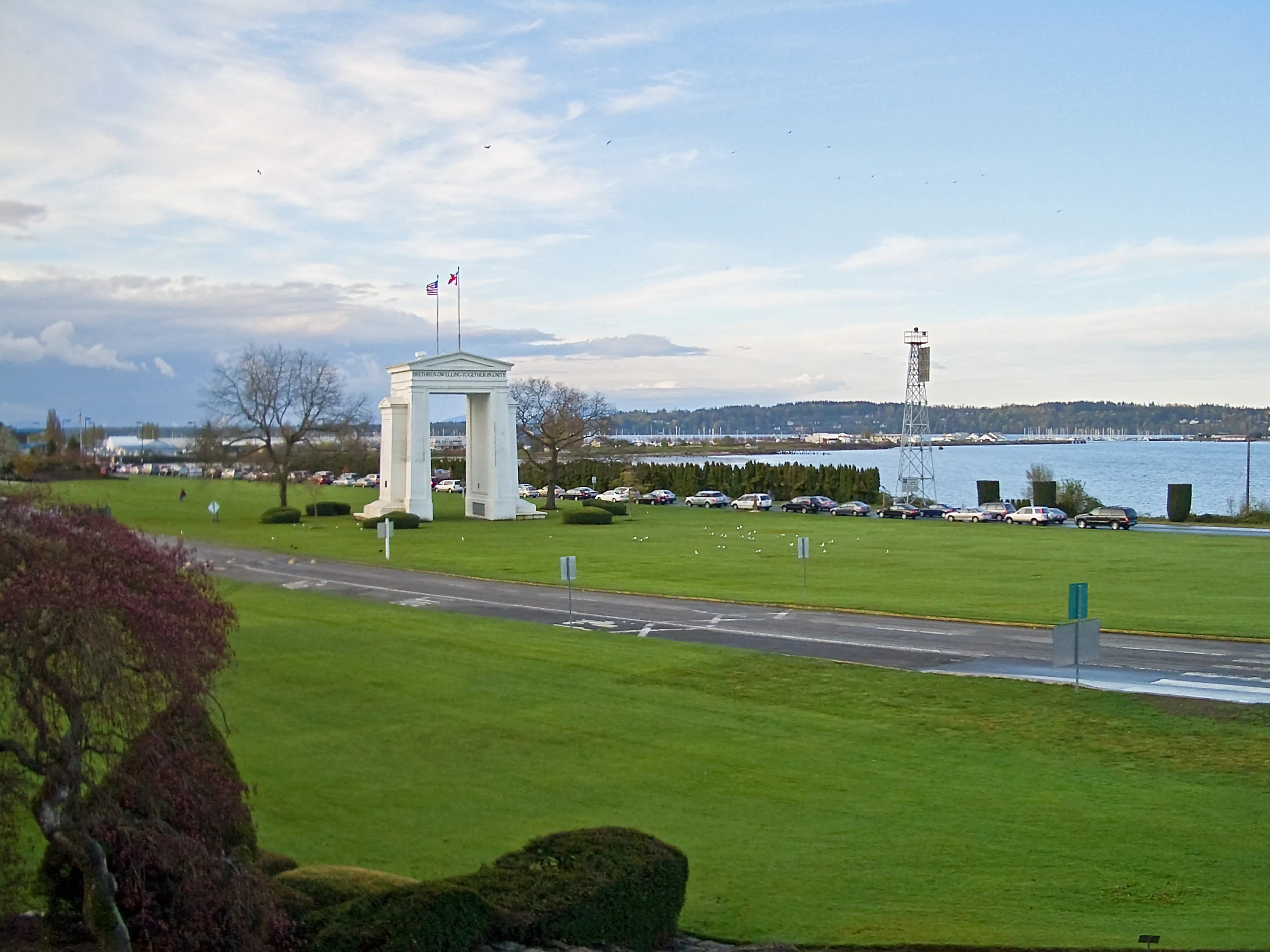 A white stone arch monument stands in a manicured green lawn near a calm bay, with cars visible on a nearby road.