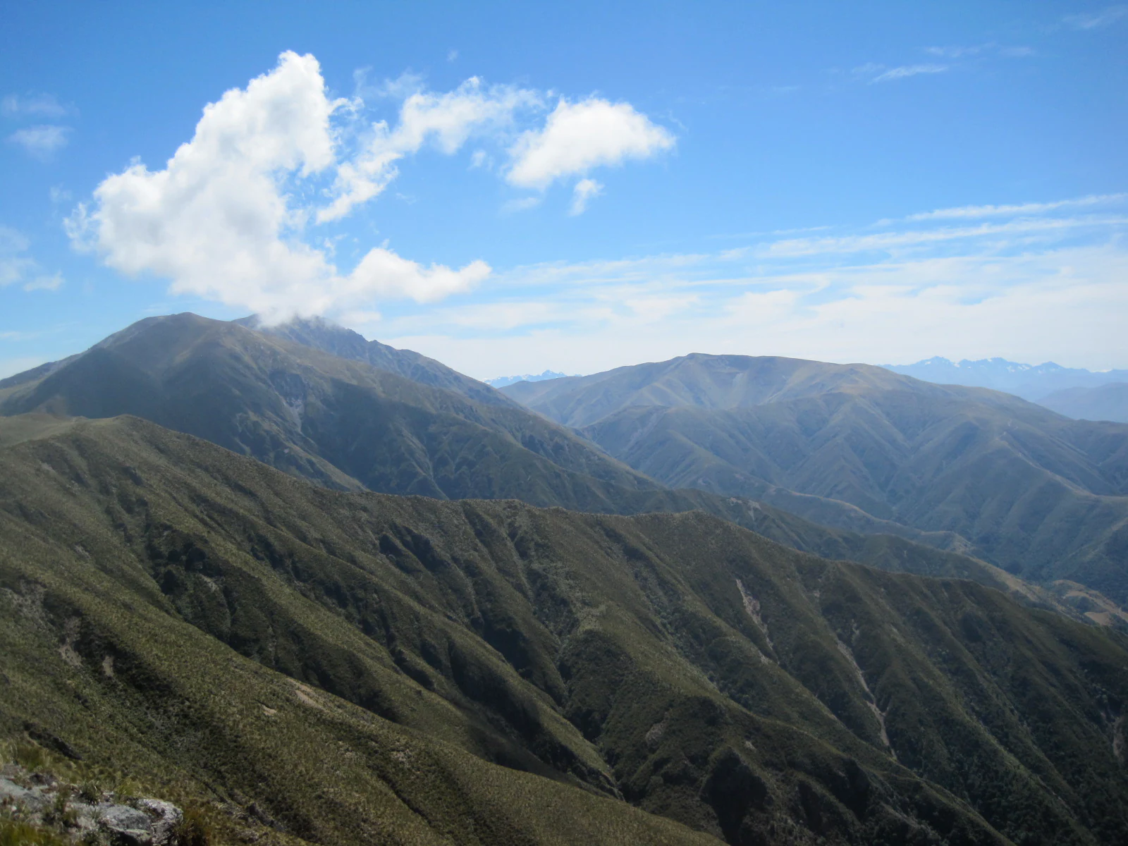 Un amplio paisaje montañoso bajo un cielo azul brillante con algunas nubes blancas, que muestra laderas montañosas onduladas cubiertas de vegetación.