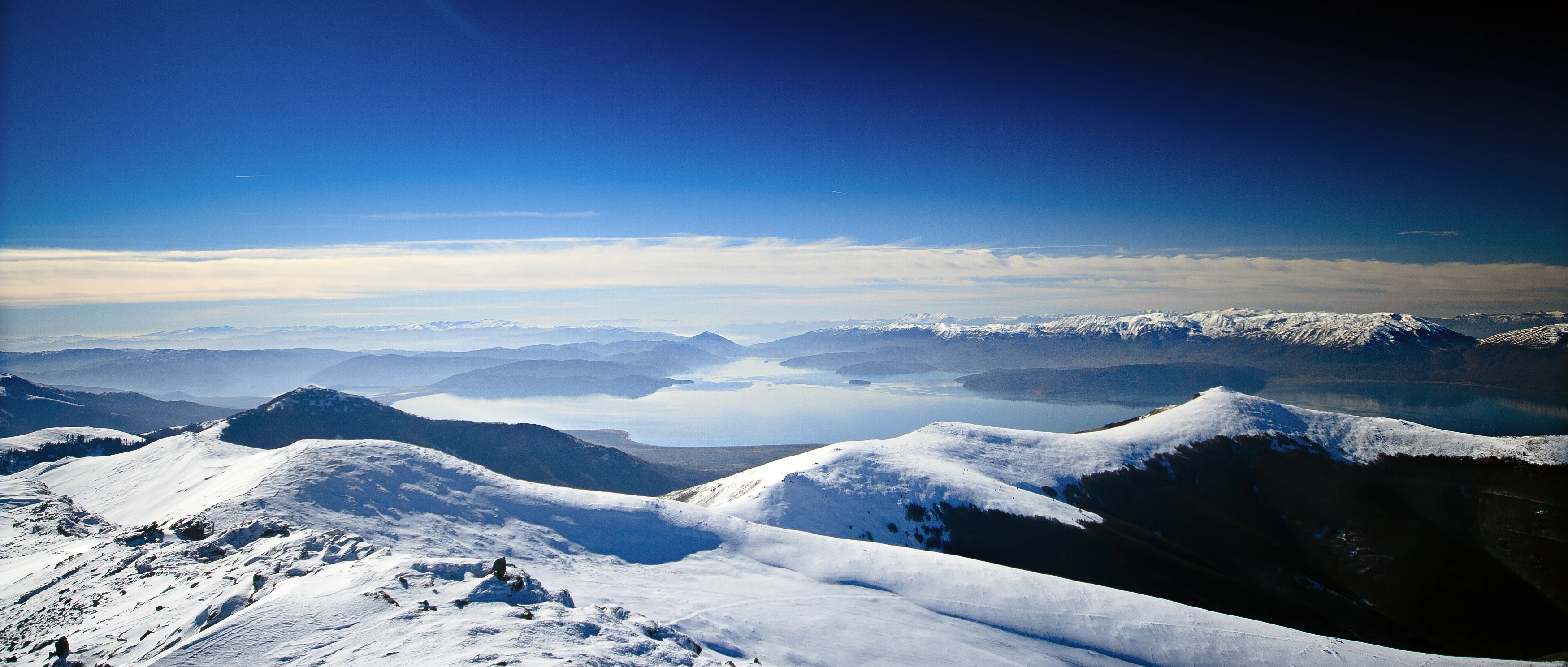 Panoramic view of snow-covered mountain peaks and Lake Prespa under a clear blue sky during winter