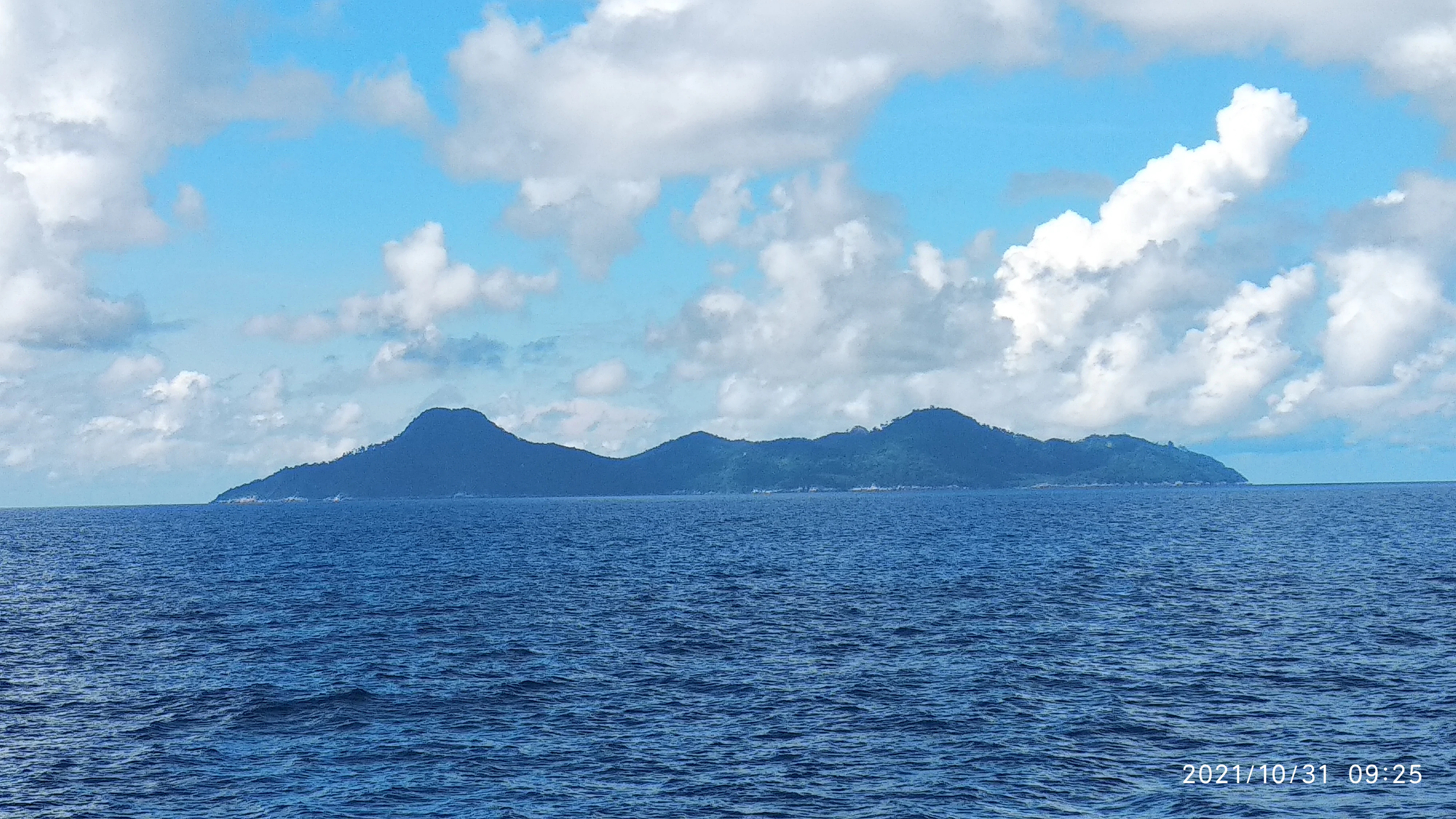 A distant view of a hilly, forested island silhouette surrounded by the open ocean under a bright, cloudy blue sky.