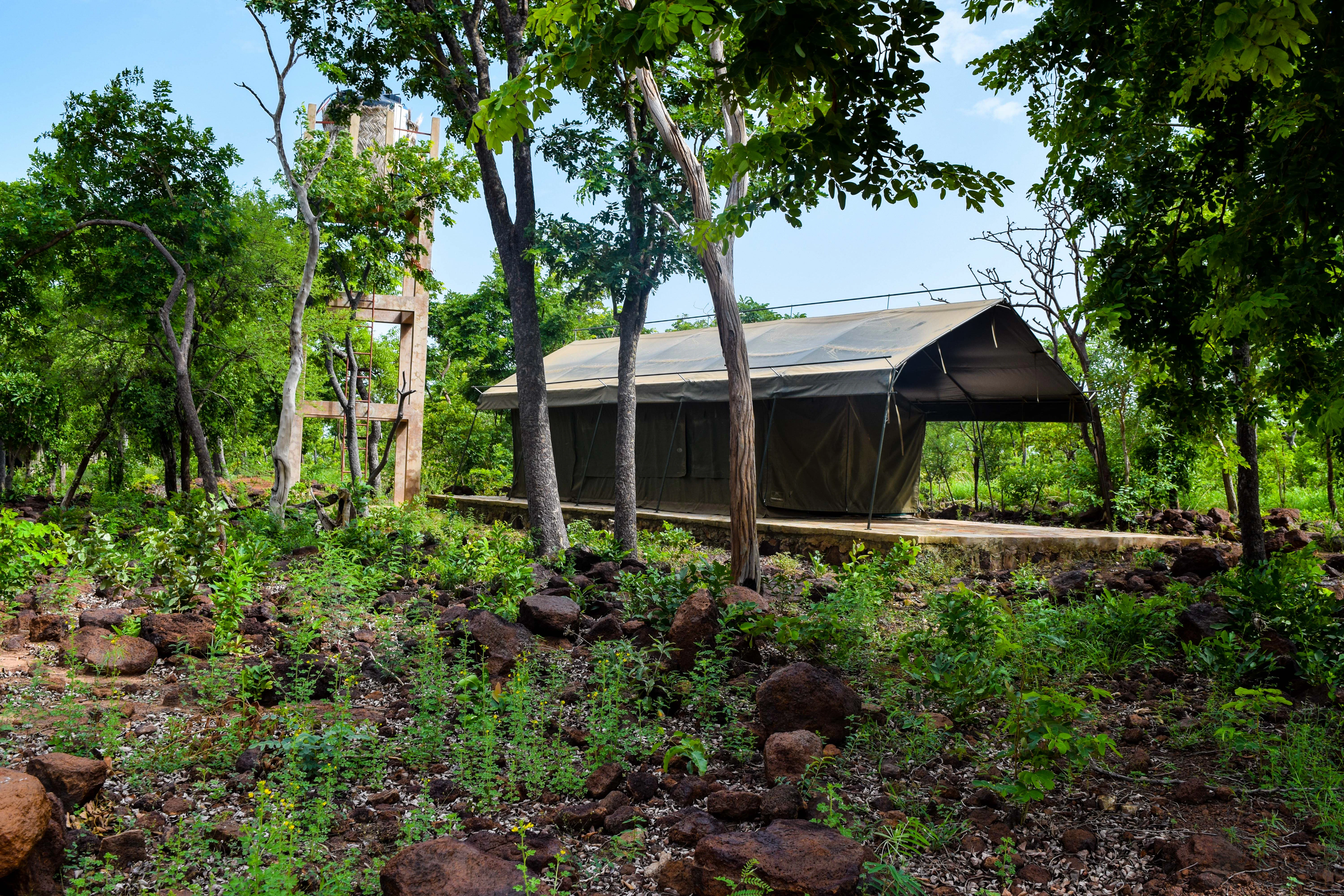 A permanent canvas safari tent on a concrete platform set amongst trees and rocky ground, with a tall, simple water tower structure visible nearby.