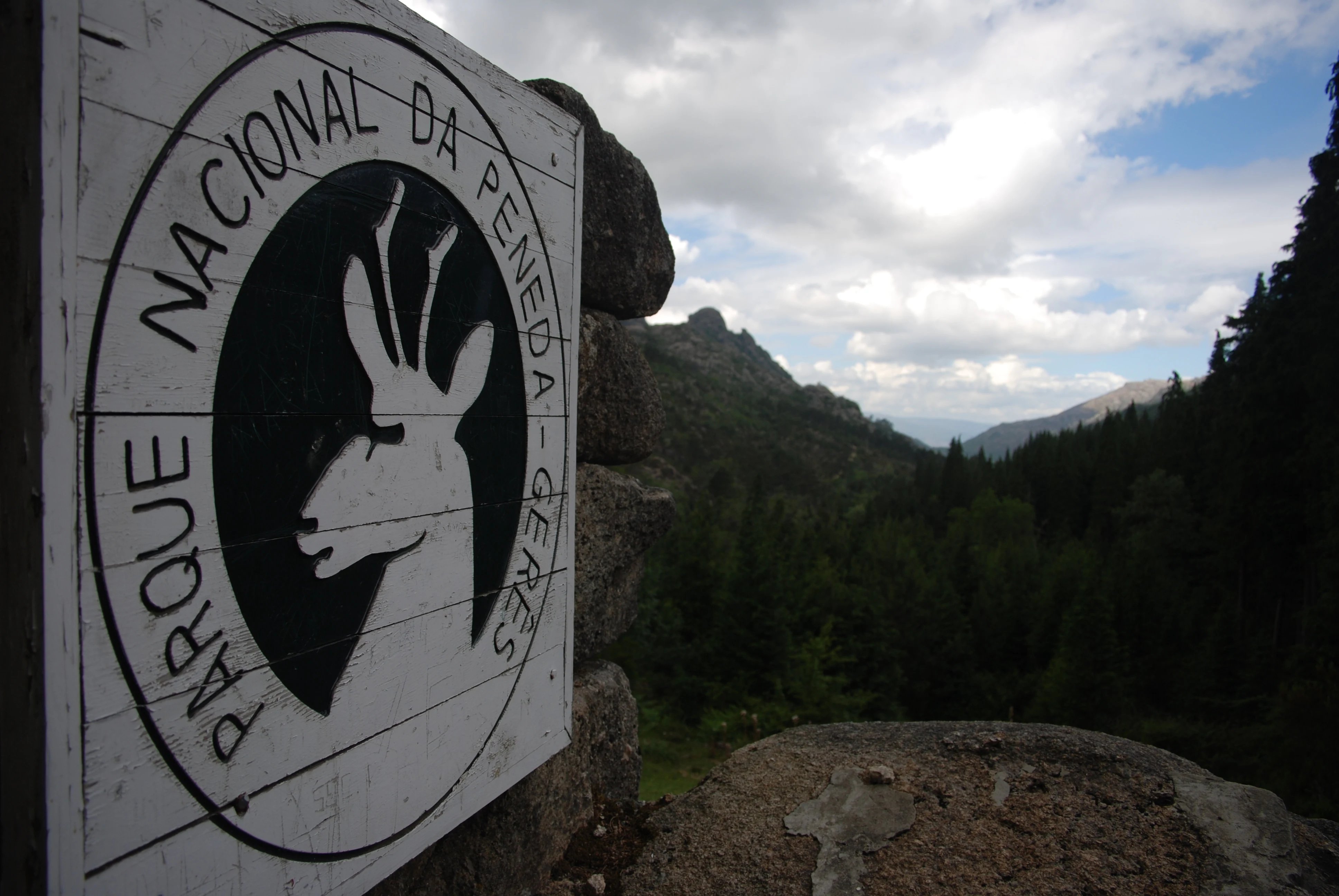 Circular park sign with text 'Parque Nacional da Peneda Gerês' and deer logo mounted on stone wall, showing forested mountains and partly cloudy sky
