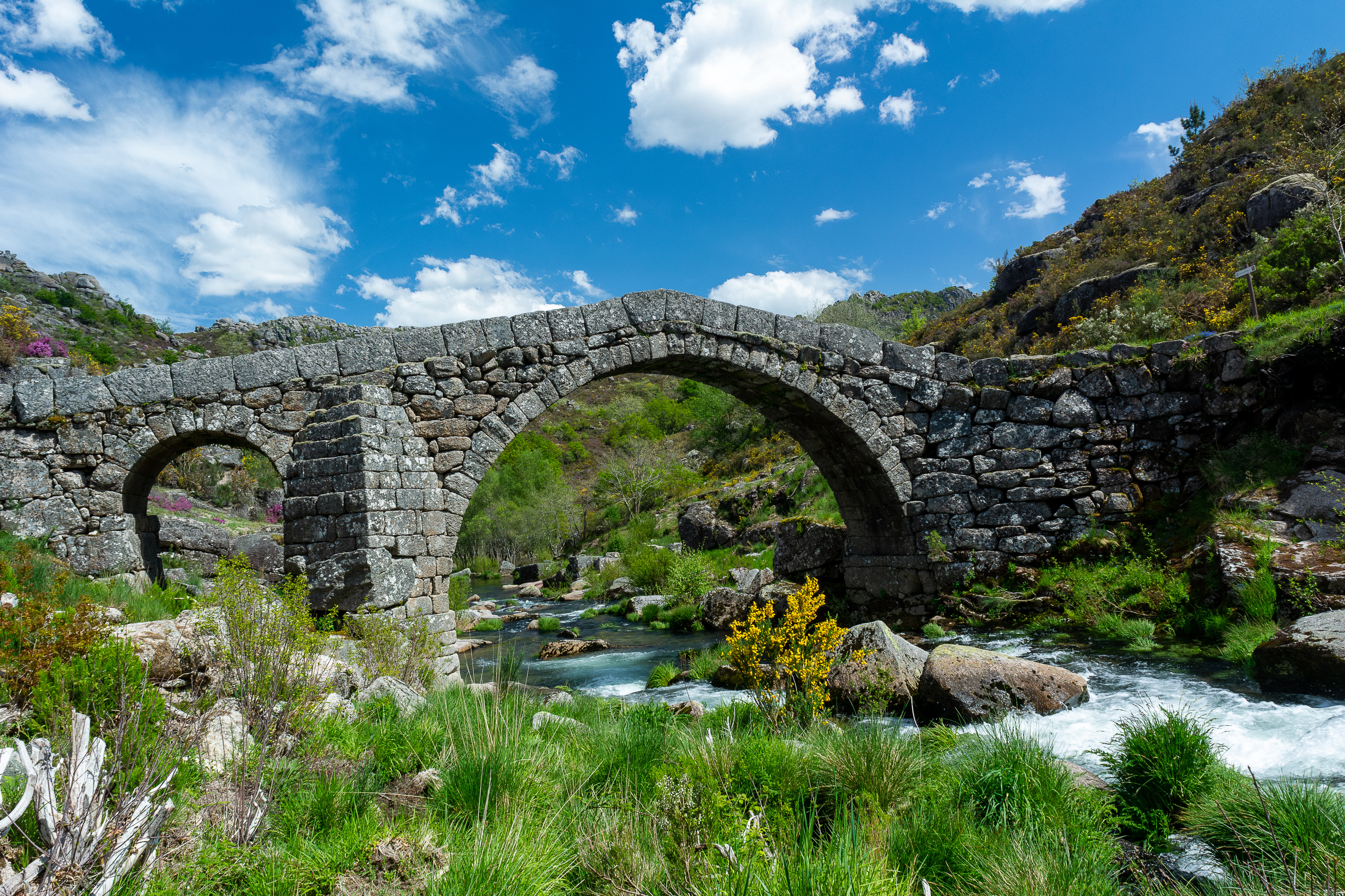 Stone arch bridge with multiple openings crossing a flowing river, surrounded by green vegetation and rocky terrain under a blue sky with white clouds