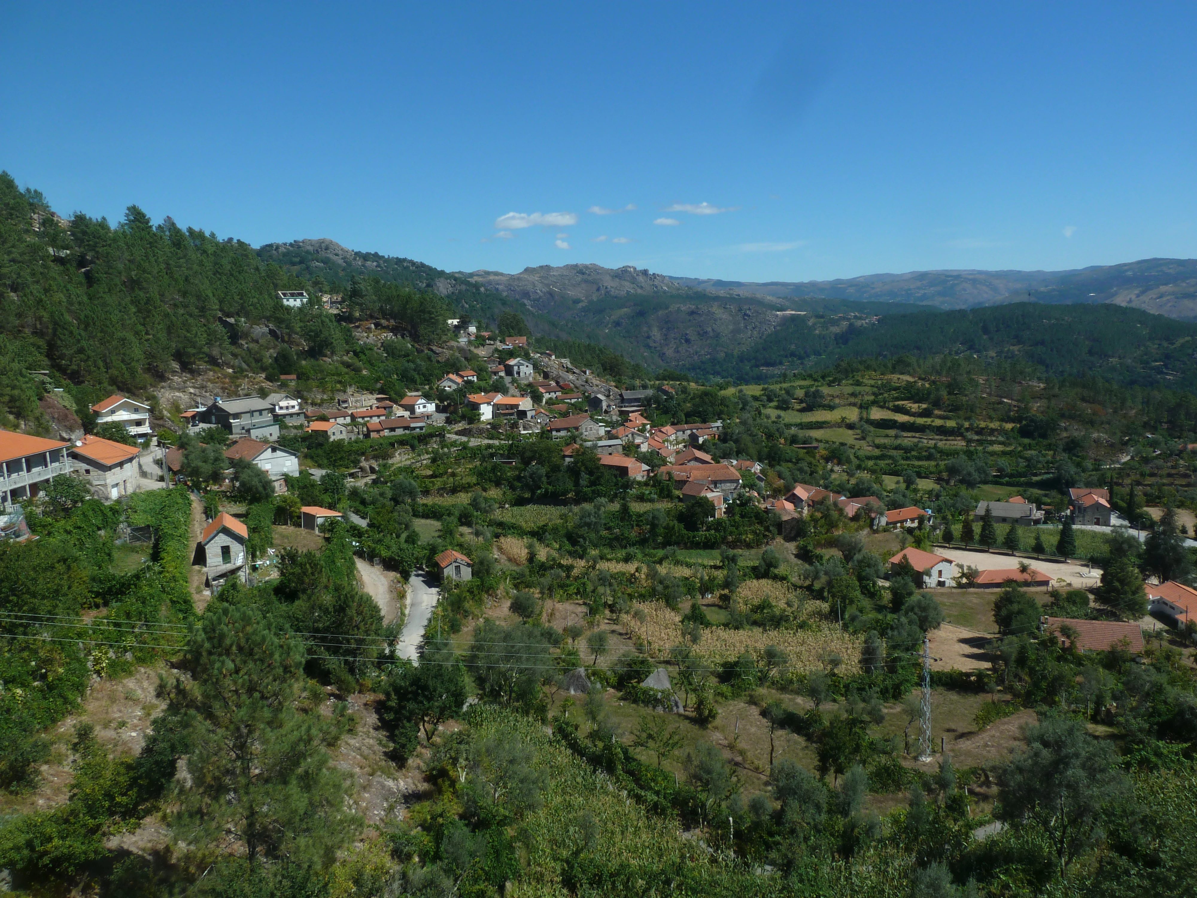 Panoramic view of a rural village with red-roofed houses, green fields, and forested hills under a clear blue sky